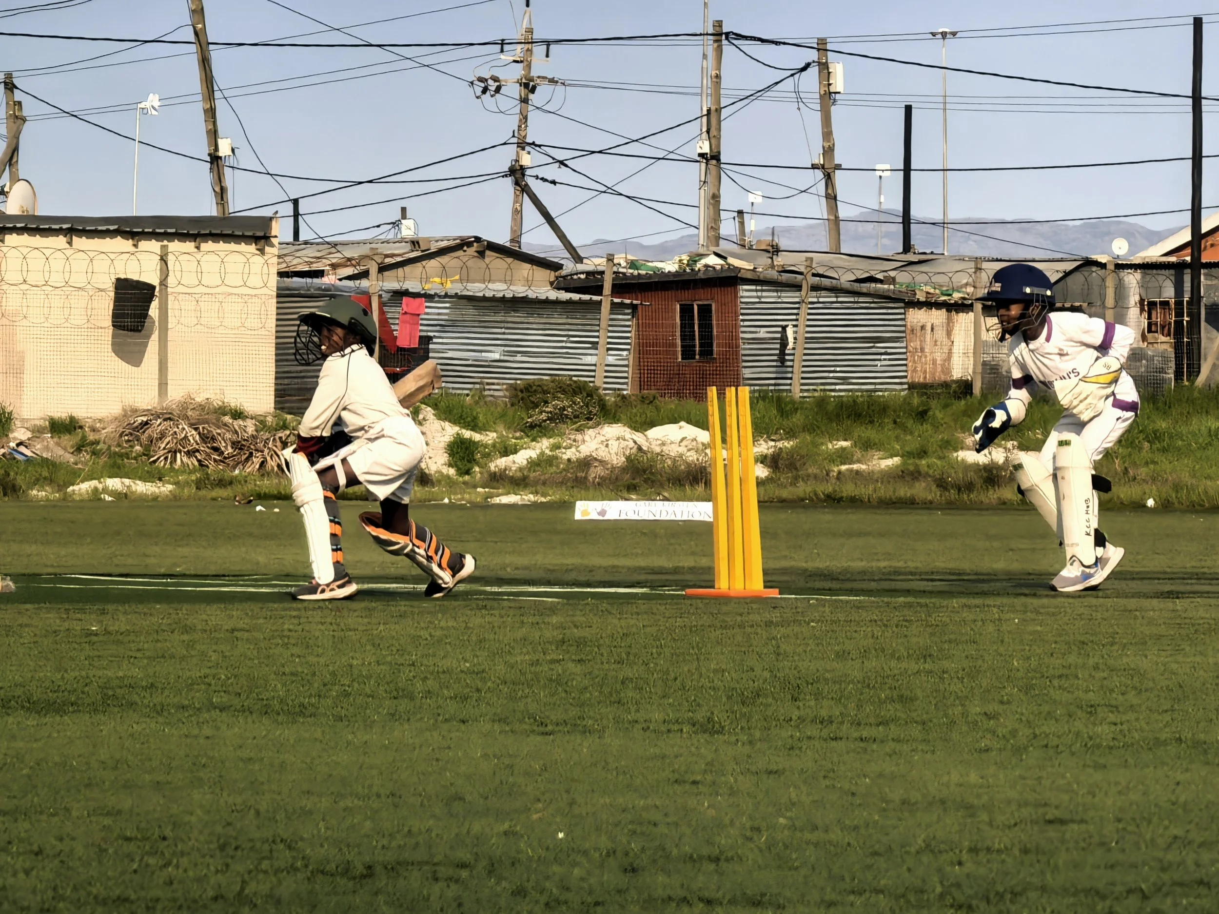 A cricket match with two players dressed in cricket gear on a grassy field, with a fence, utility poles, and buildings in the background.