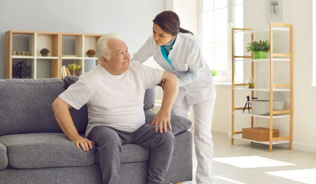A healthcare worker assists an elderly man sitting on a gray couch during a home visit in a well-lit living room.