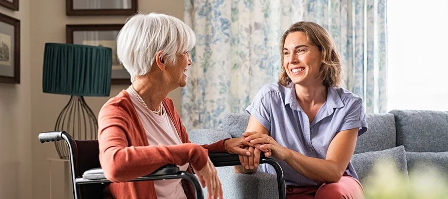 A young woman with short brown hair smiling and holding hands with an elderly woman with white hair sitting in a wheelchair in a bright living room.