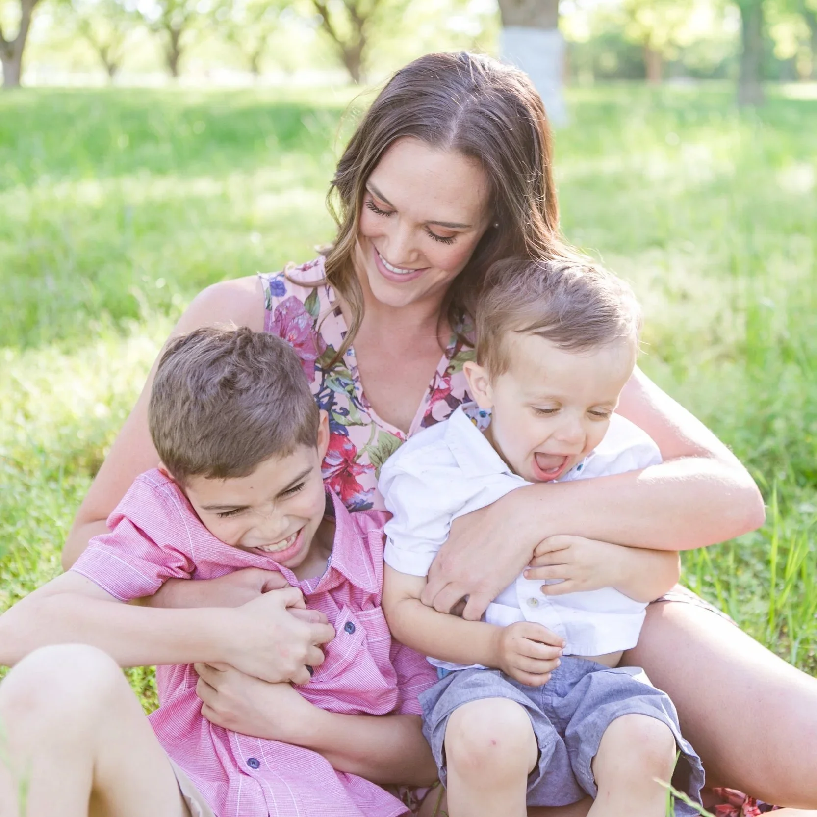 A woman with two young boys sitting on the grass and playing together outdoors on a sunny day.