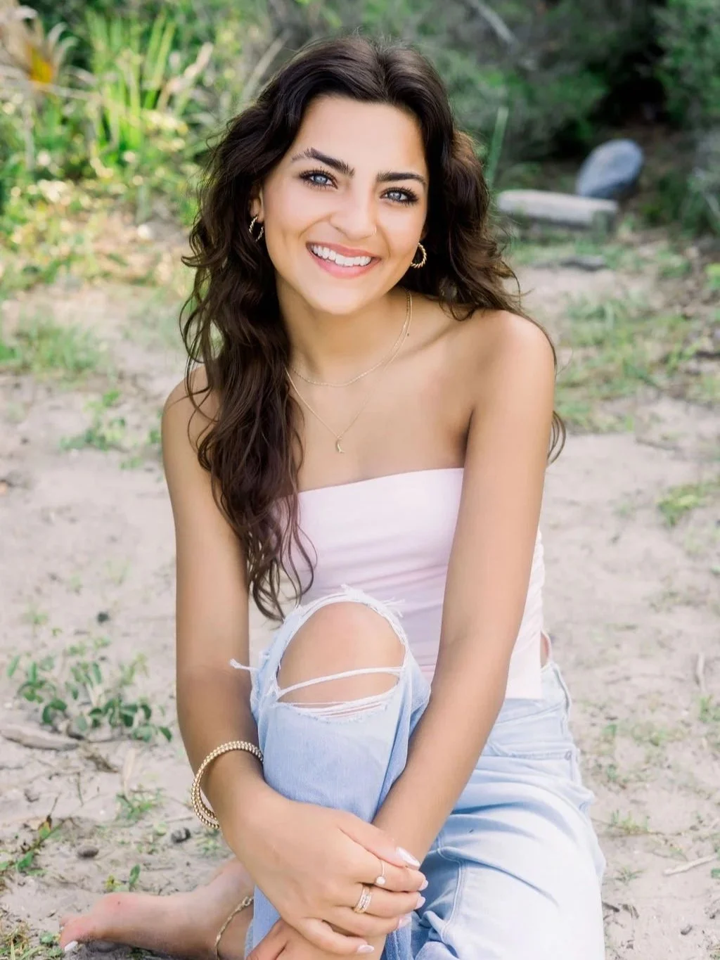 senior brunette girl sitting on beach in a light pink top and ripped jeans