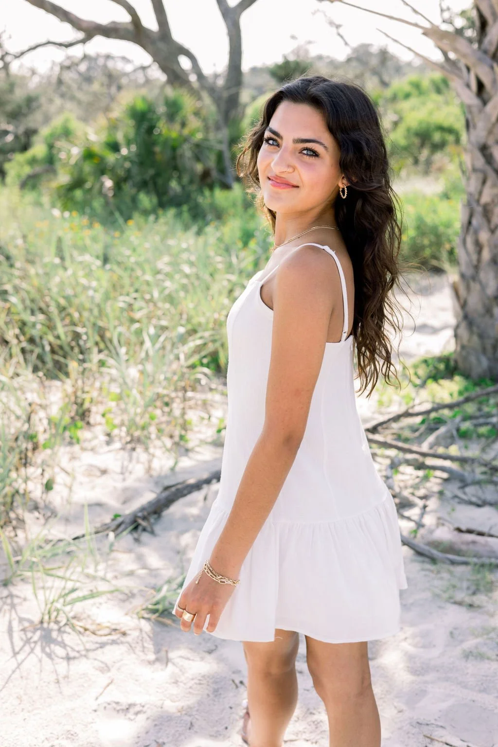 brunette senior in a white dress looking back smiling while on the beach