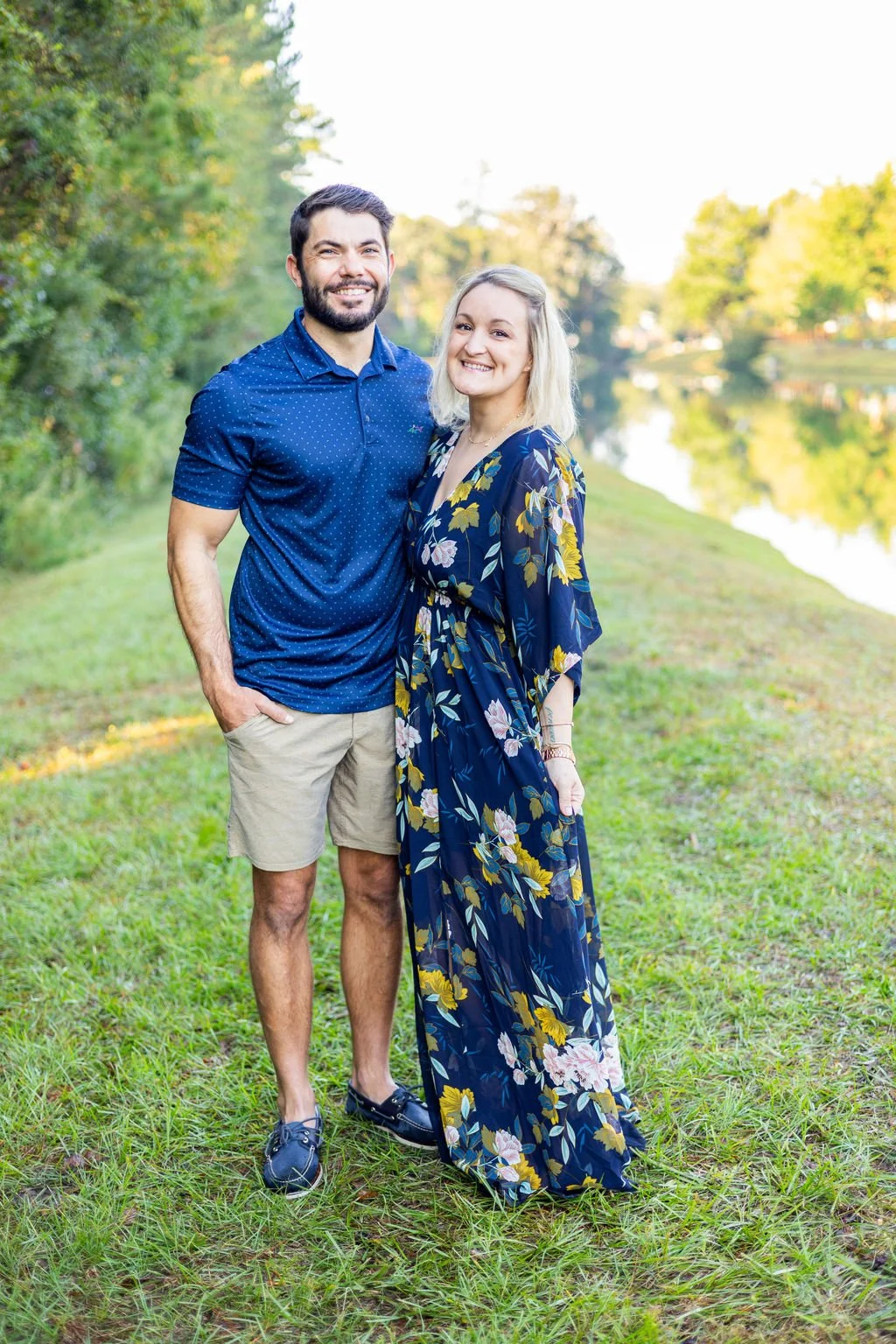 A smiling couple standing outdoors near a river or lake, with trees and green grass in the background during daytime.