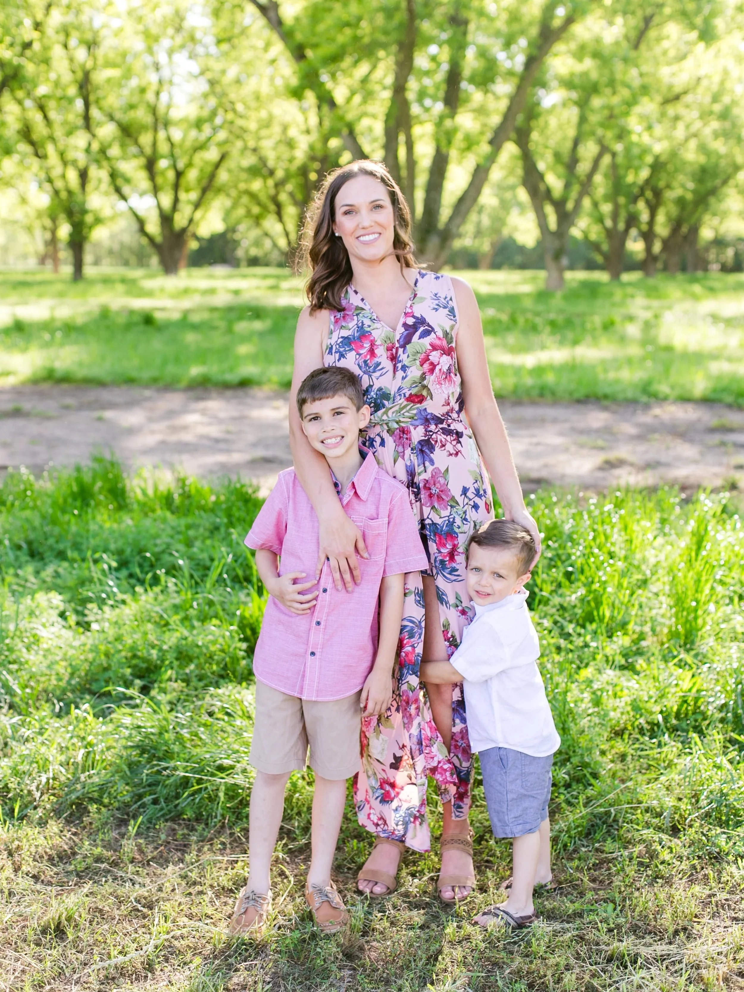 mother in pink floral dress holding her two sons in a tree farm
