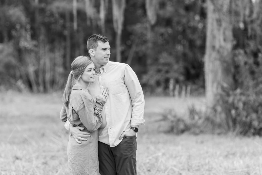 A young couple stands closely together outside in a natural setting, with trees and grass in the background, smiling and looking at the distance.
