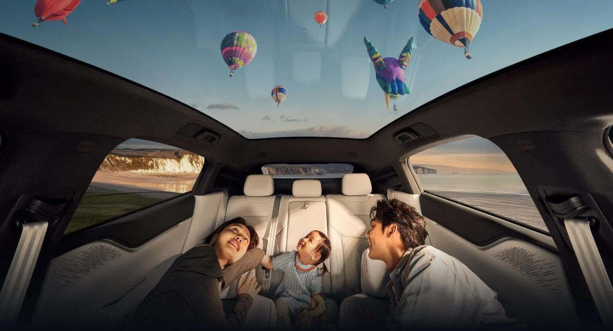 Une famille de trois personnes assise dans l'arrière d'une voiture, regardant par le toit panoramique un ciel rempli de montgolfières colorées au coucher du soleil.