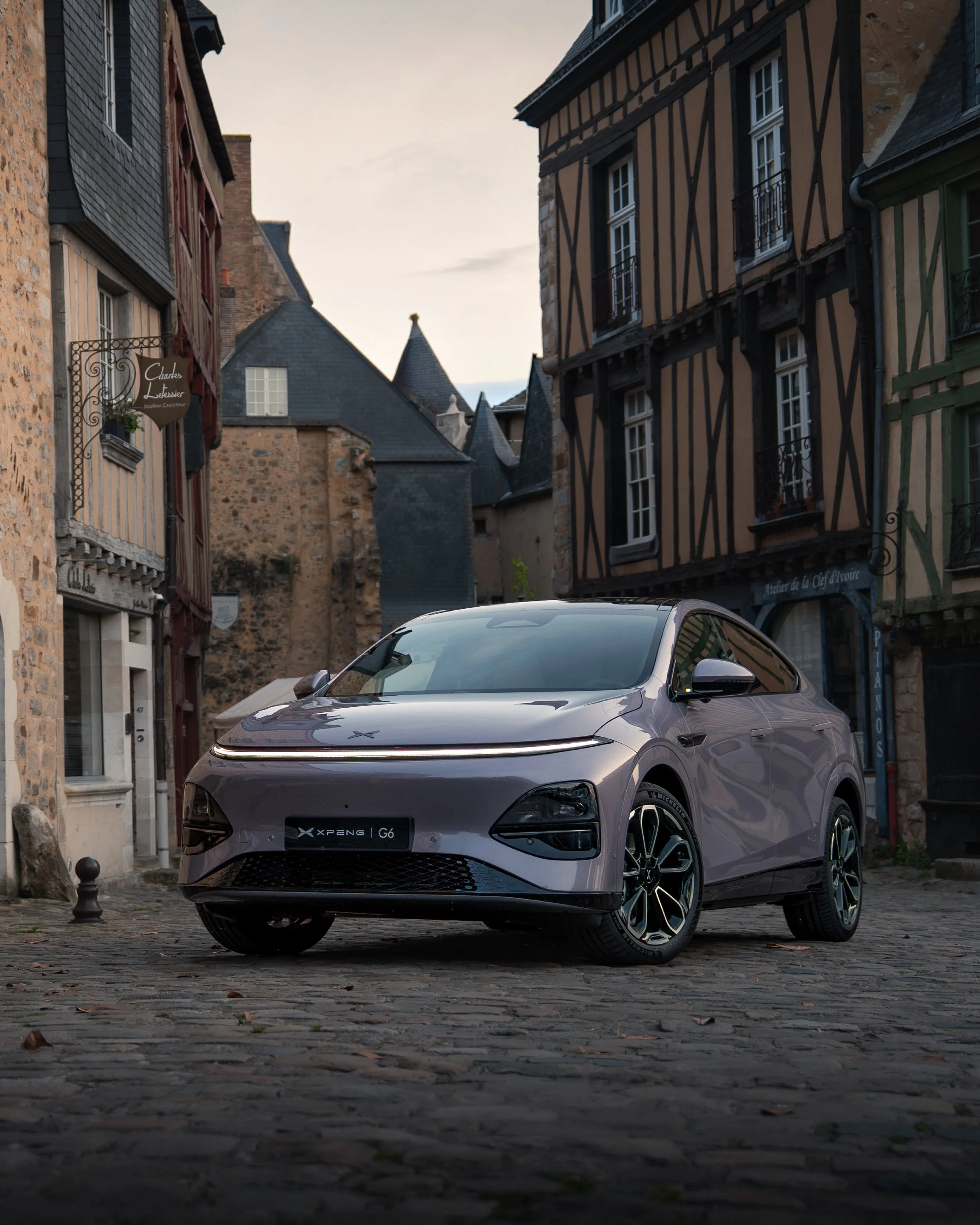 Voiture électrique grise stationnée sur une rue pavée dans un vieux quartier avec des bâtiments en pierre et en bois.