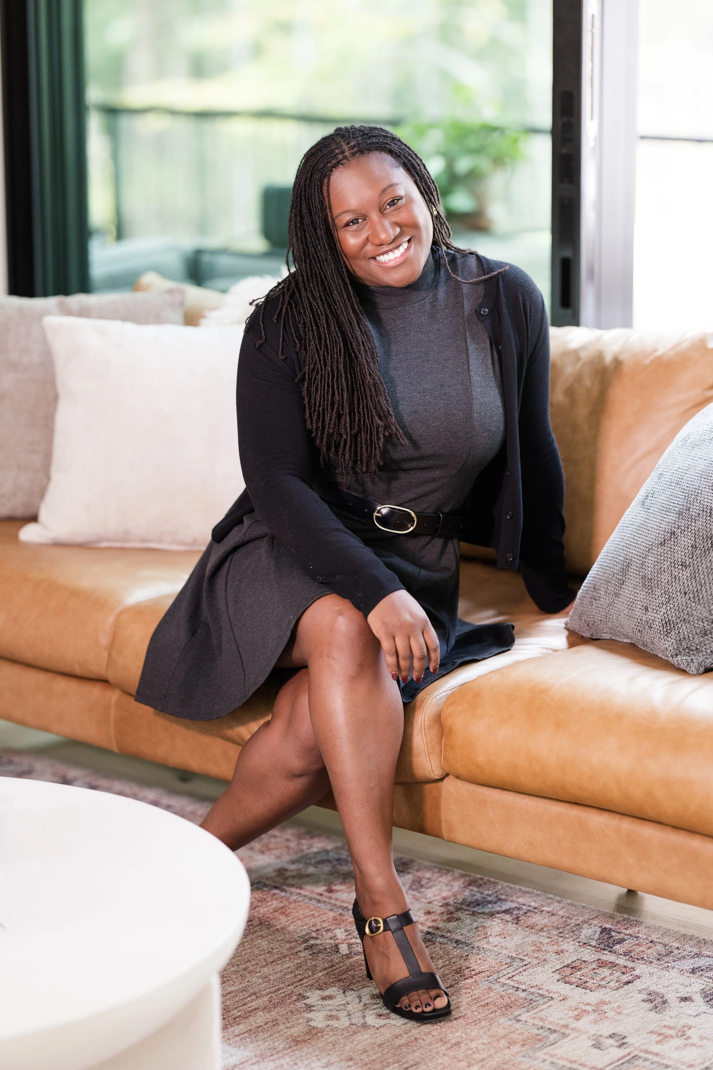A woman with long braided hair smiling while sitting on a tan leather sofa in a bright living room with large windows, decorated with throw pillows and a rug.