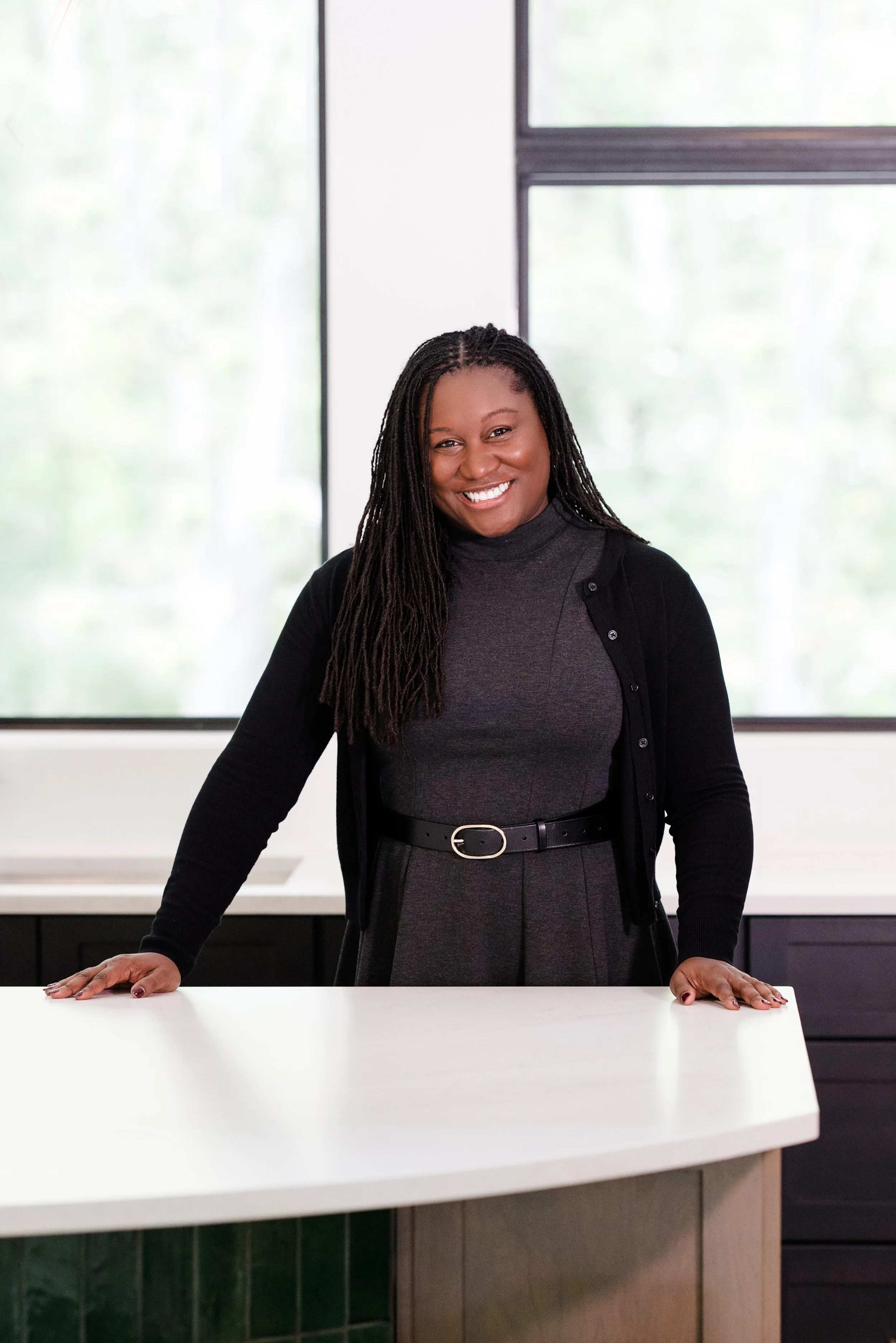 A woman with long braided hair, wearing a gray dress with a black belt, smiling and leaning on a white table in a bright room with large windows.