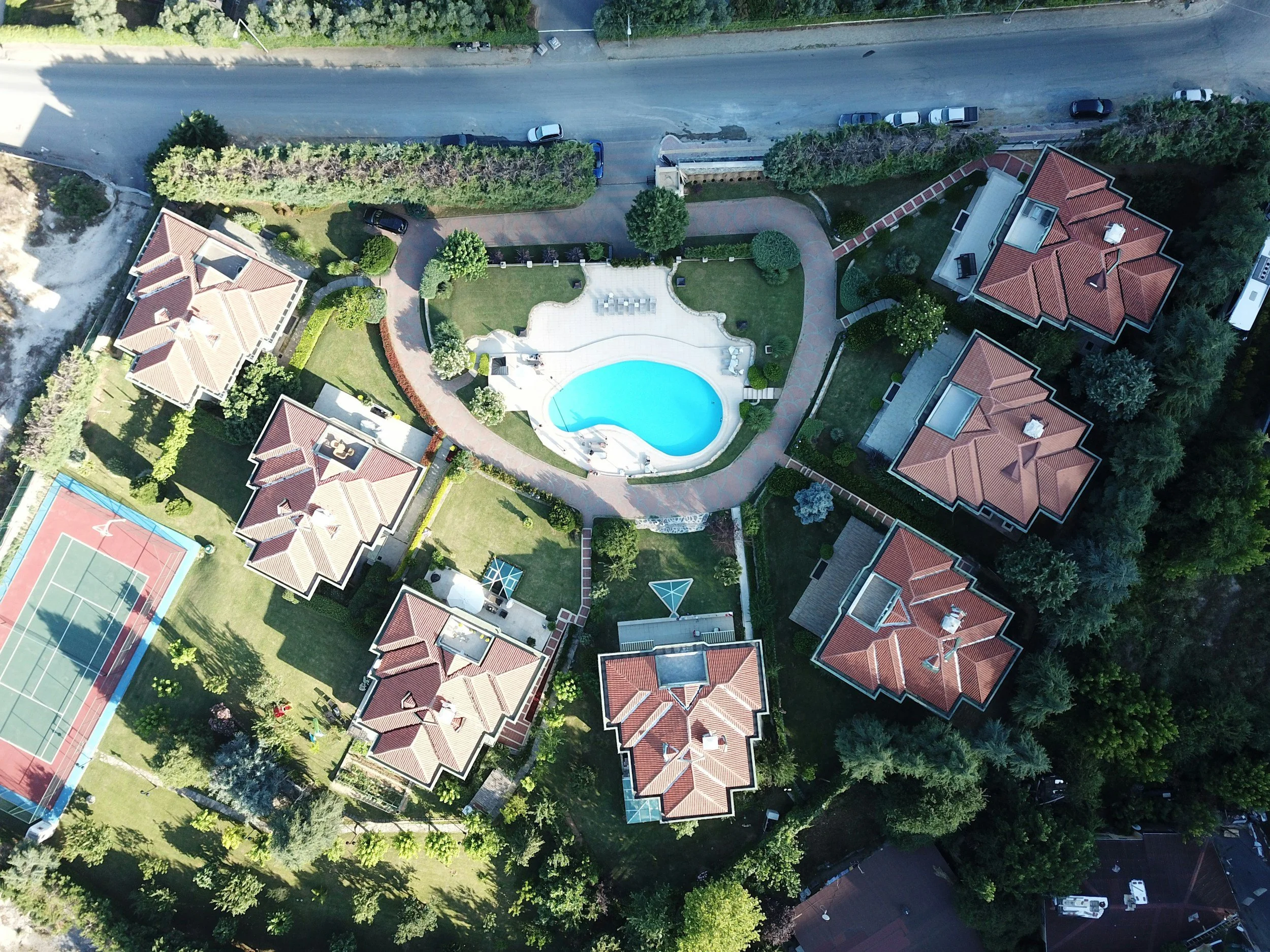 An aerial view of a residential complex with multiple houses featuring red-tiled roofs, a central swimming pool, a tennis court, and surrounding green lawns and trees.
