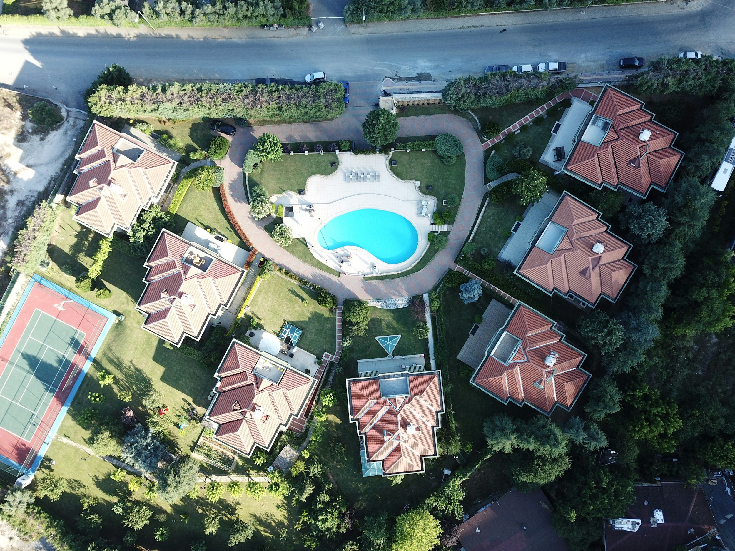 Aerial view of a residential area showing multiple houses with red-tiled roofs, a swimming pool, a tennis court, and greenery.