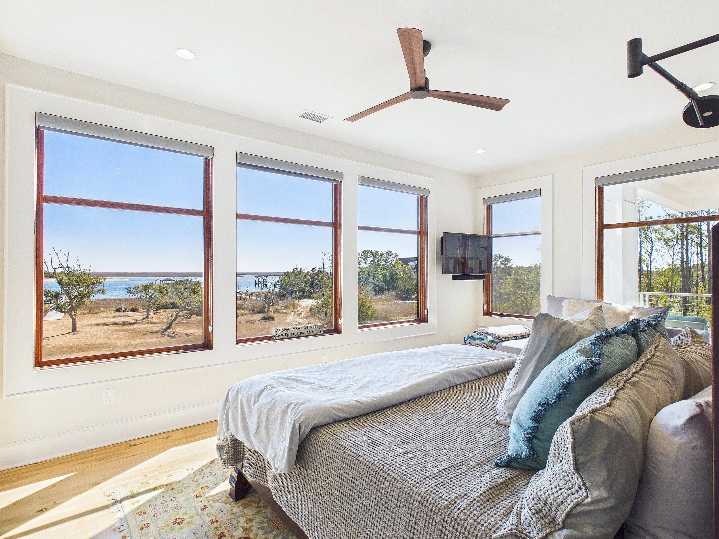 Bright bedroom with large windows overlooking a beach and trees, with a bed, multiple pillows, a wall-mounted TV, ceiling fan, and wooden floors.