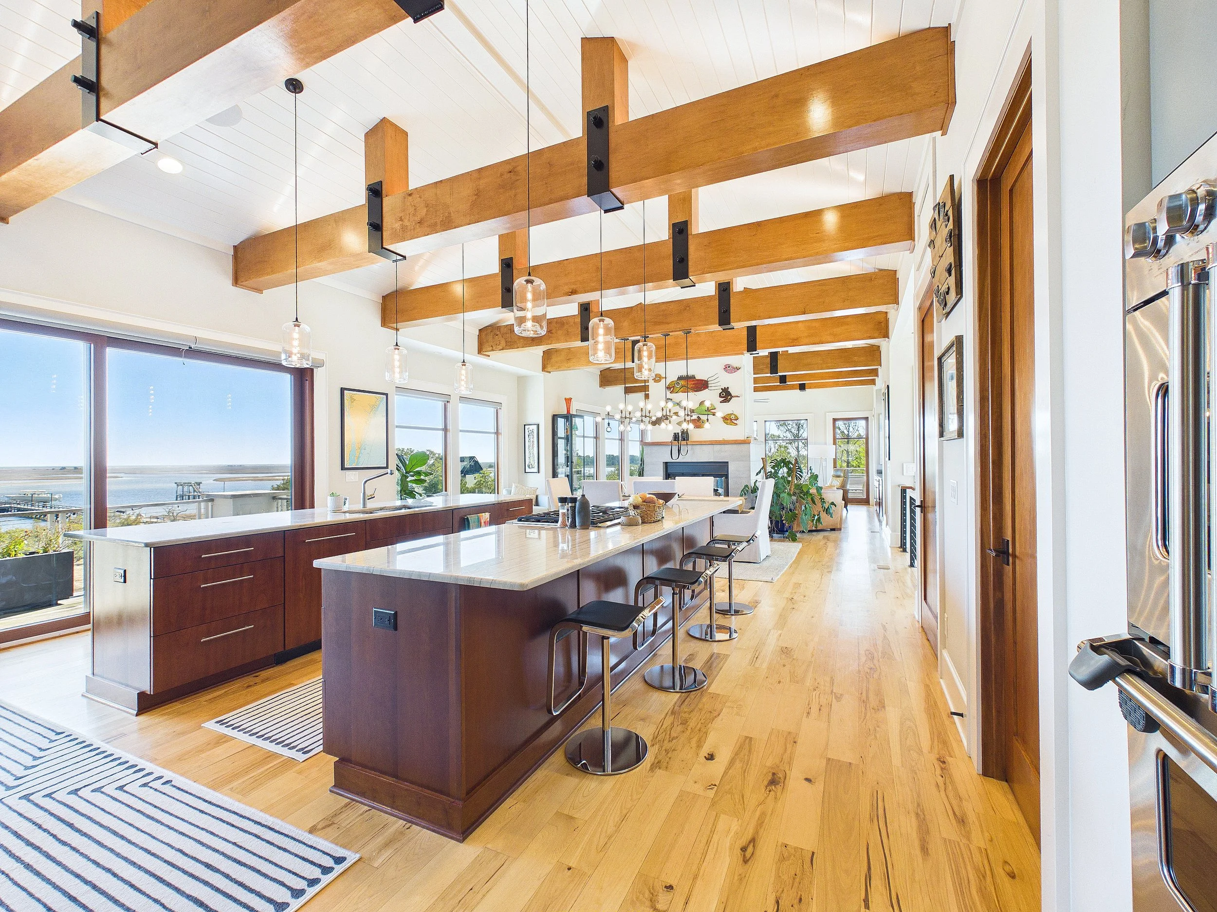 Open-concept kitchen and living space with wooden beams and flooring, large windows, island countertop with barstools, and casual dining area.