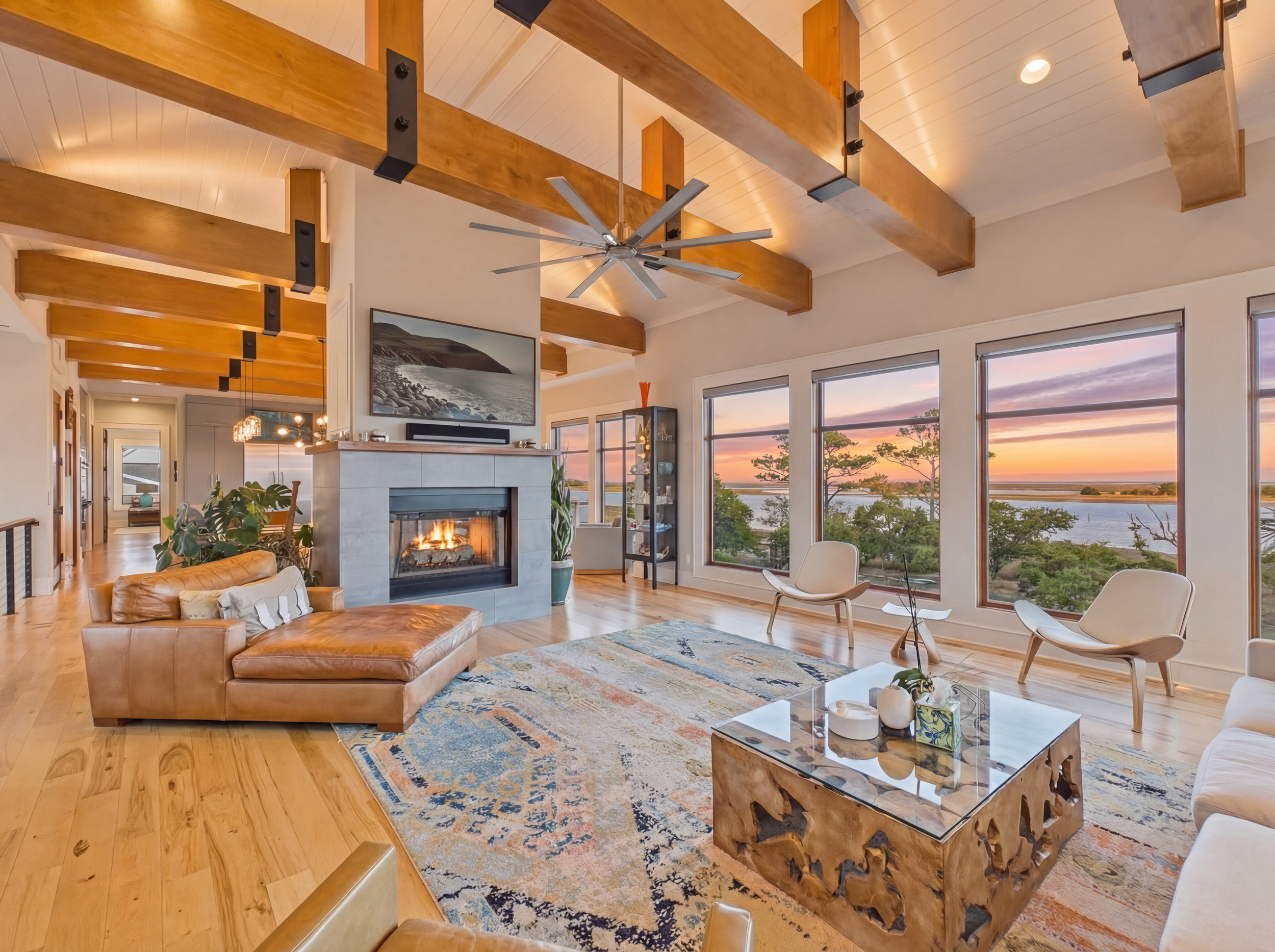Bright living room with large windows overlooking a lake, featuring a tan leather sofa, white armchairs, a colorful area rug, and a modern coffee table, with wooden beams on the ceiling and a fireplace.