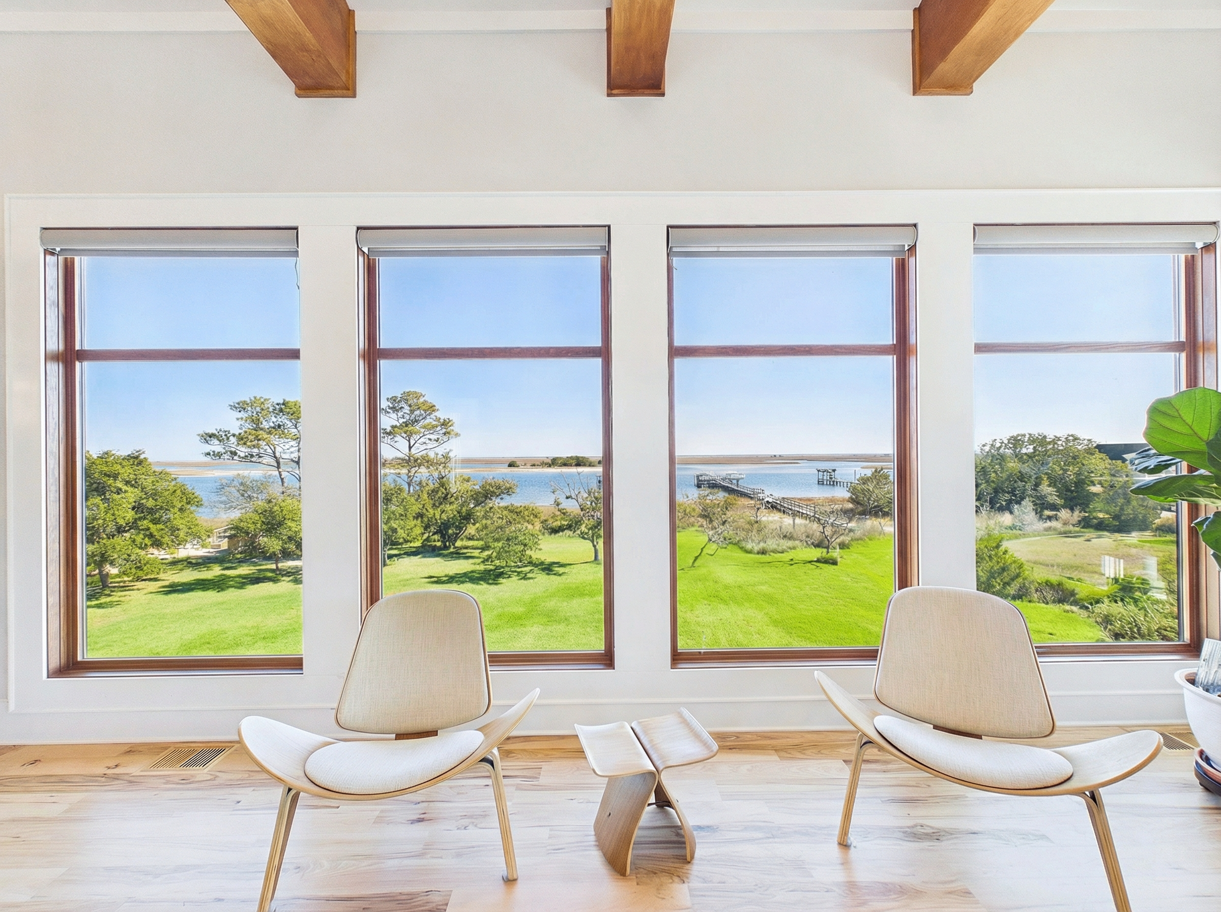 Living room with large windows overlooking a beach, two modern white chairs, a small wooden table, and a potted plant.