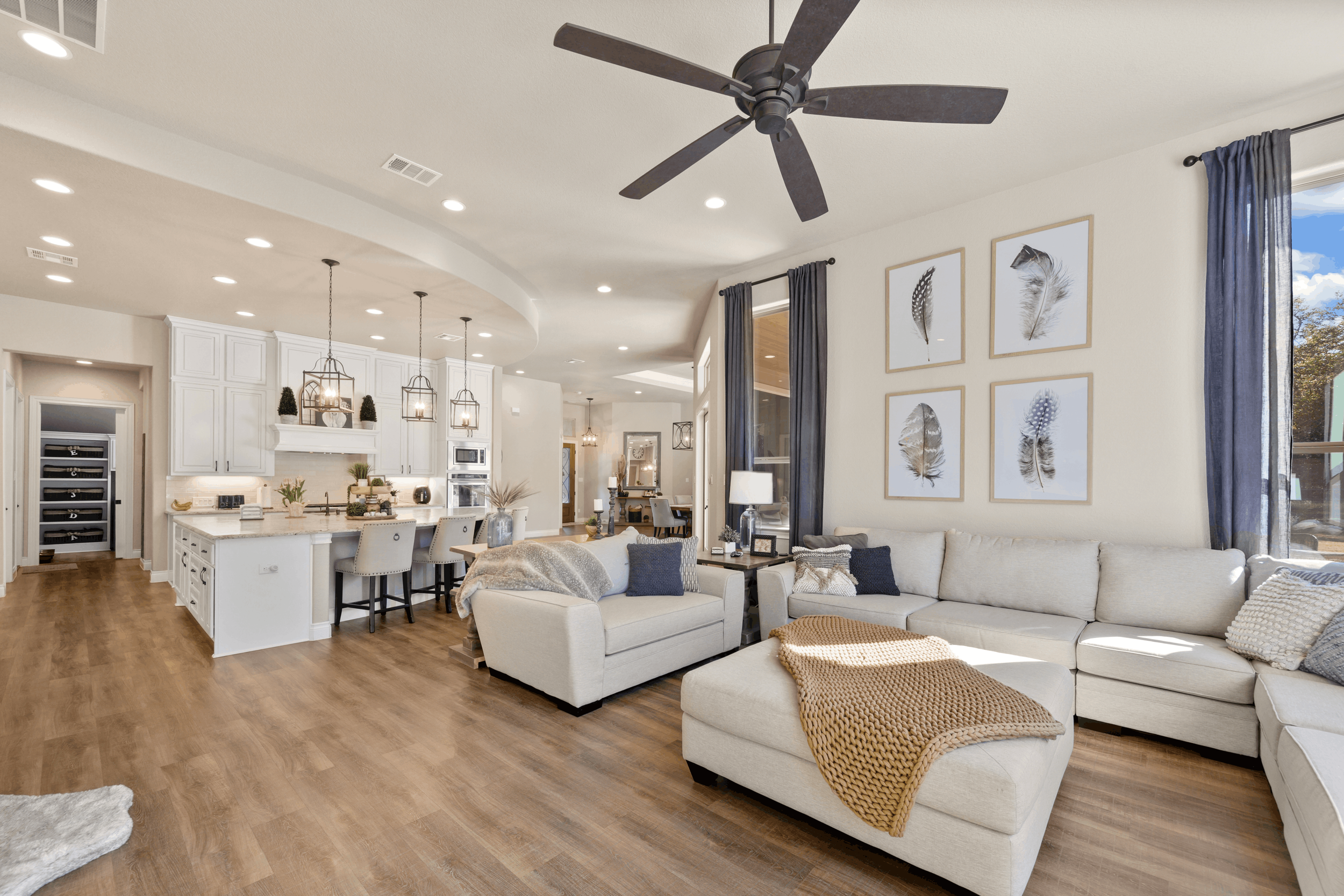 Living room with a large white sectional sofa, a beige armchair, framed feather artwork on the wall, dark blue curtains, and a ceiling fan. Open kitchen in background with white cabinets and pendant lighting.
