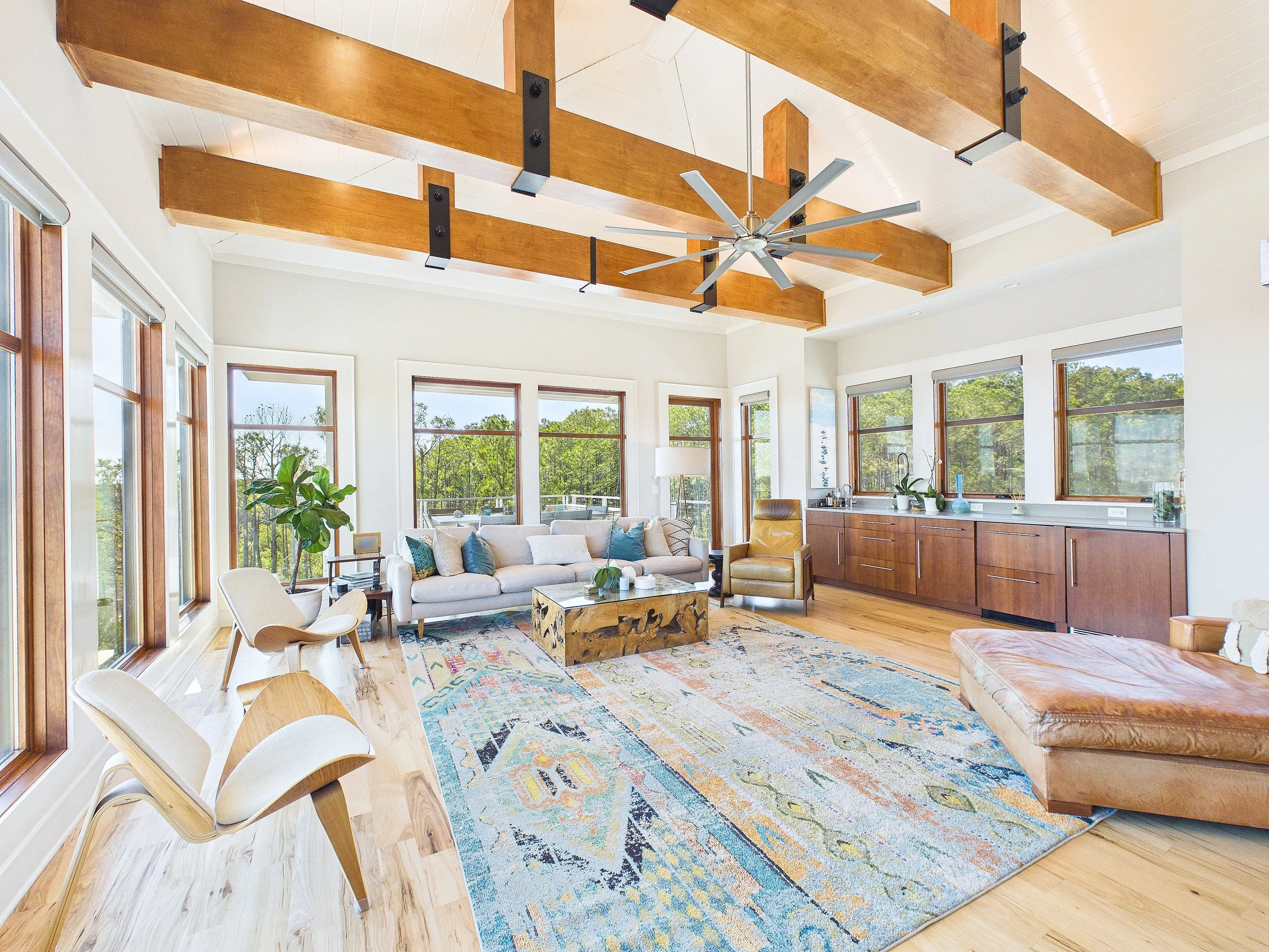 Bright living room with large windows, wooden beams on ceiling, white walls, wooden floor, and modern furniture including a white sofa, yellow armchair, leather ottoman, and a colorful rug.