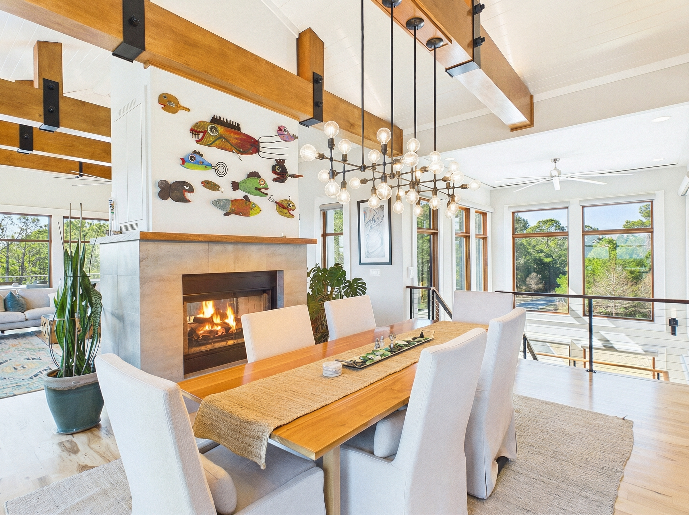 Modern dining room with a wooden table, white chairs, a fireplace, colorful wall art, chandelier, large windows showing trees, and a beige rug.
