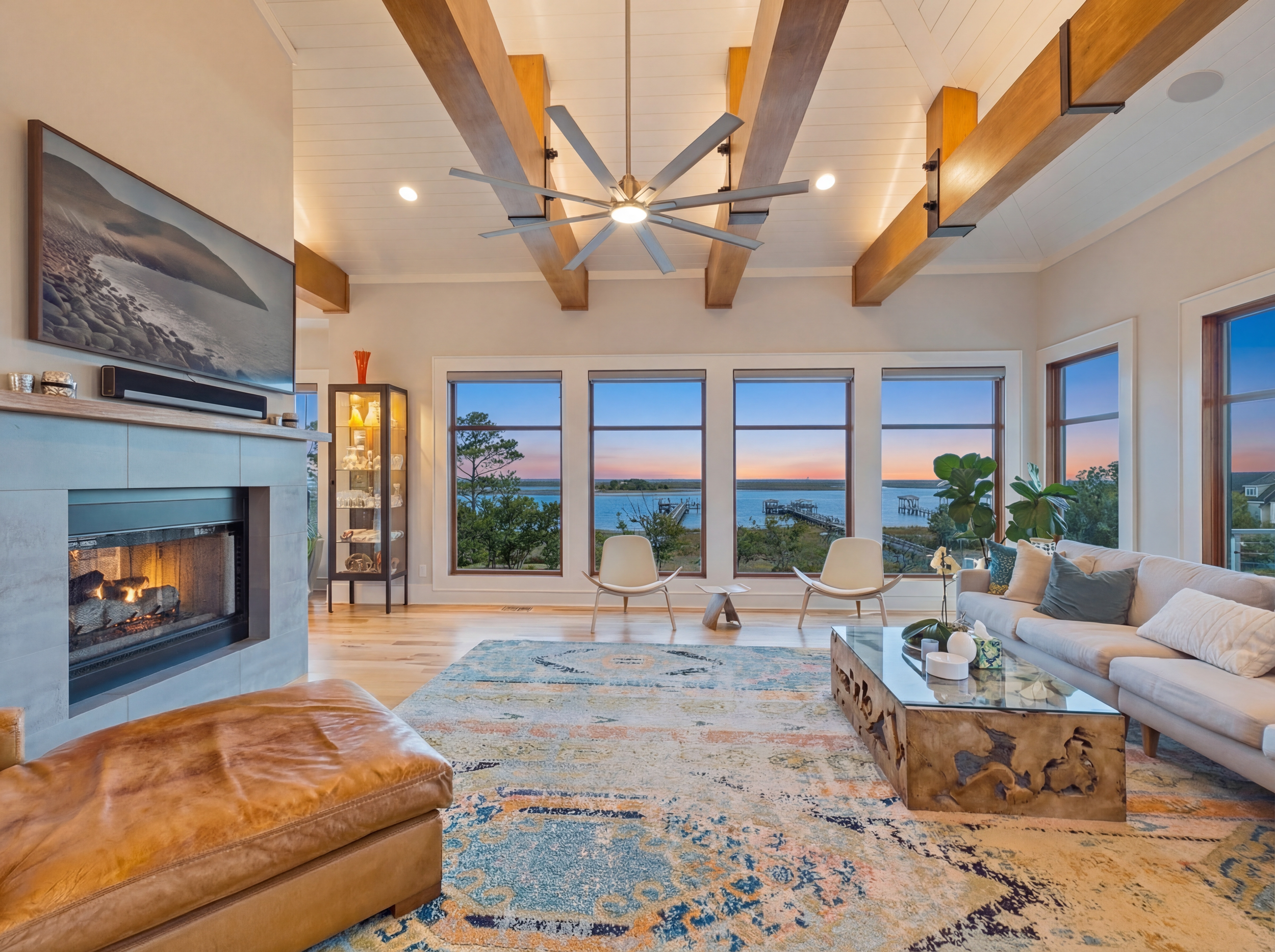 Bright living room with large windows overlooking a water view, featuring a white sofa, a wooden coffee table, a leather ottoman, a colorful area rug, and a modern ceiling fan.