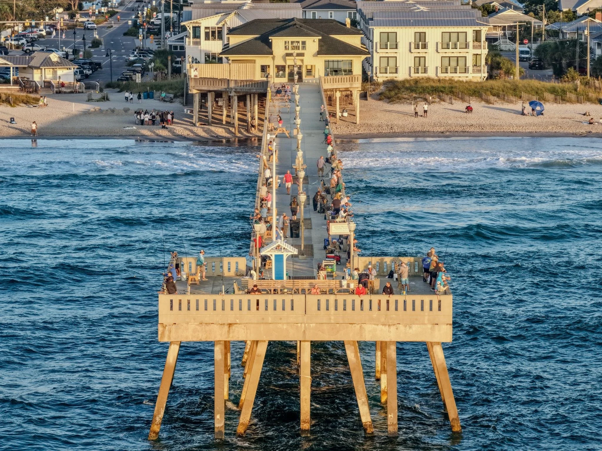 A long pier extending into the ocean with many people fishing and walking. The pier leads to a beach with houses and buildings nearby, and the water has gentle waves.