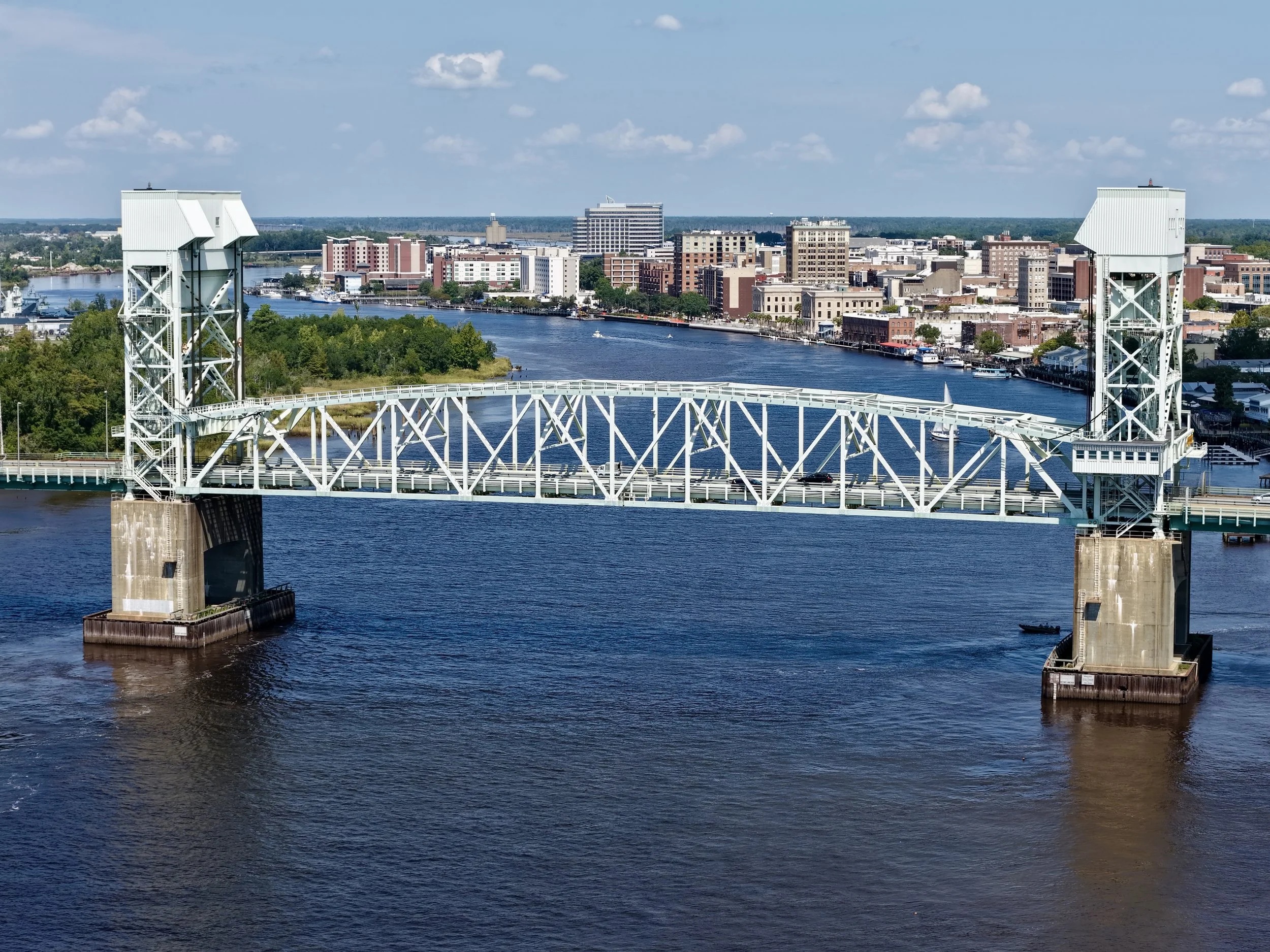 An aerial view of a large river with a bridge crossing over it and a city skyline in the background.