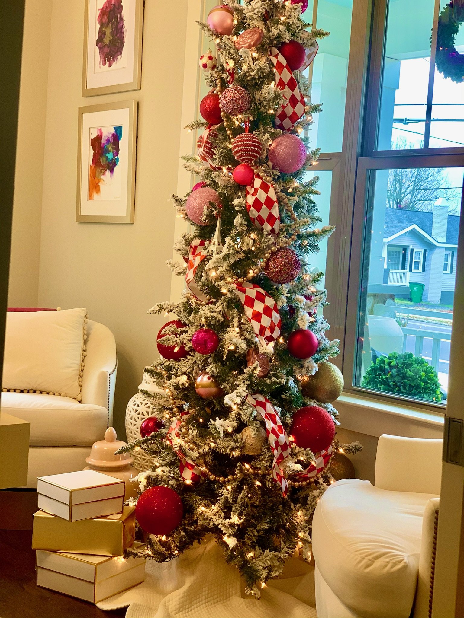 Decorated Christmas tree with red, pink, and gold ornaments, red and white checkered ribbons, and white lights, placed indoors near a window.