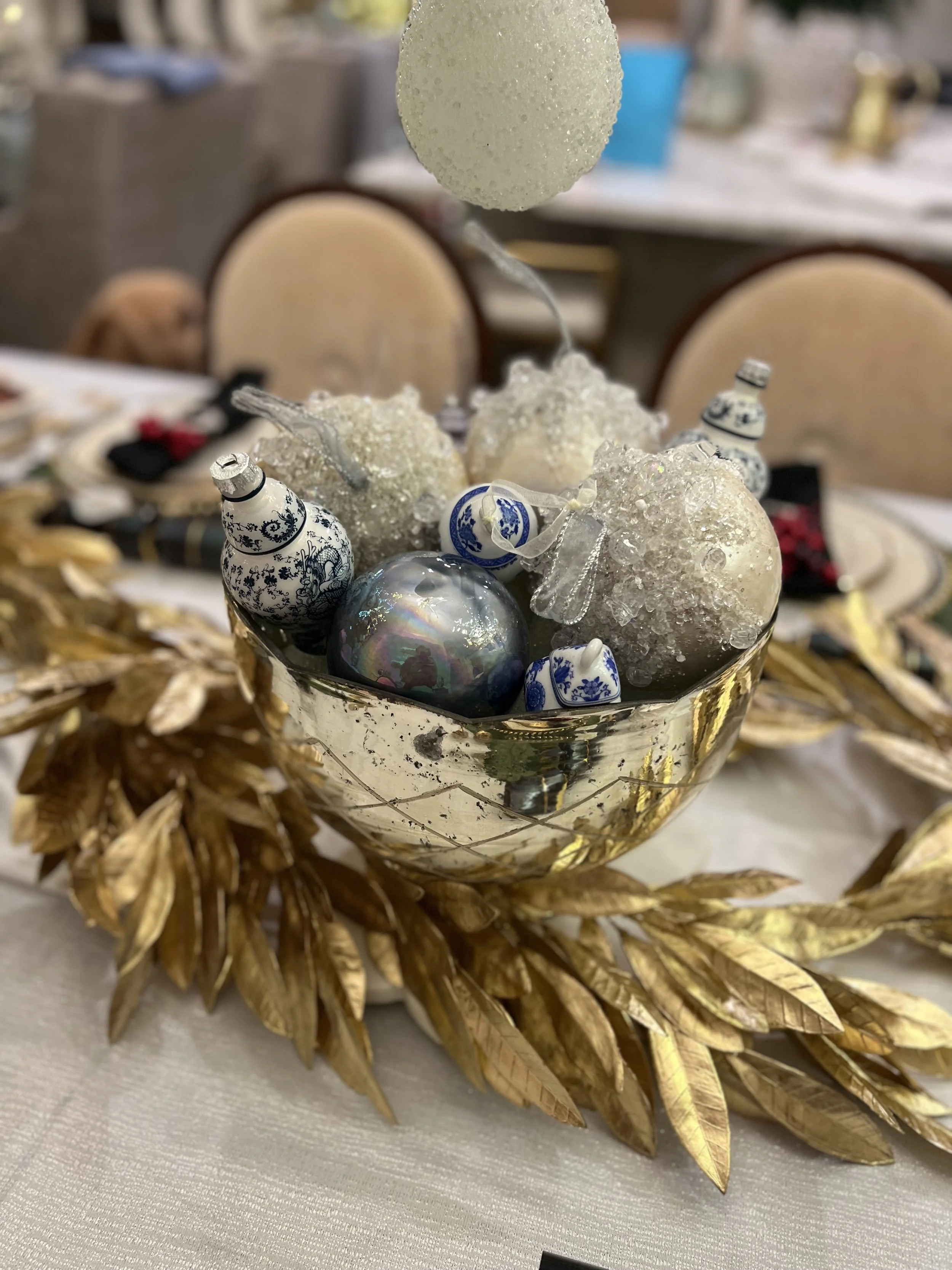 Decorative holiday centerpiece with a glass bowl filled with Christmas ornaments and frosted decorations, surrounded by gold-colored leaves arranged in a wreath-like shape.