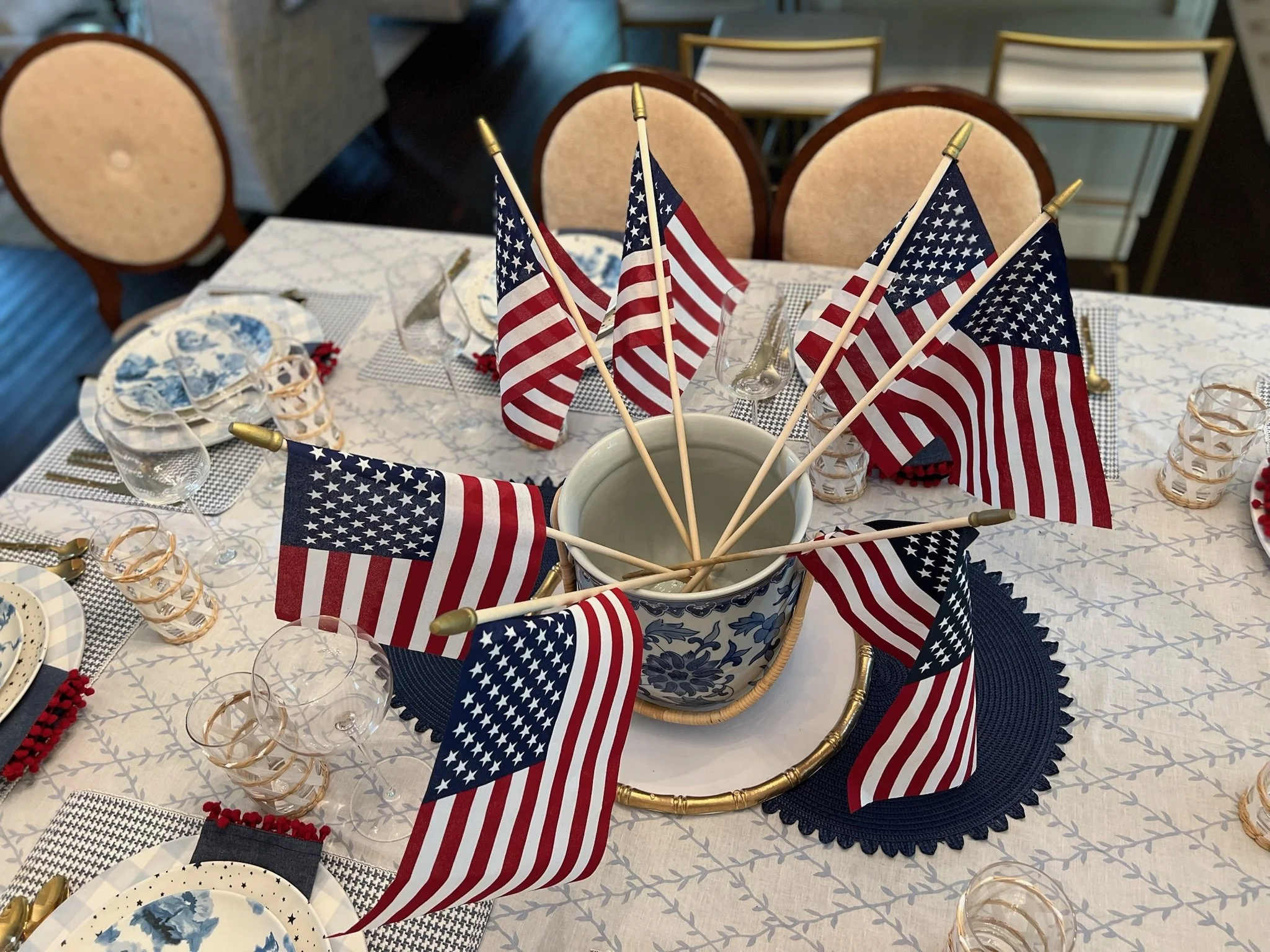 Table decorated for a patriotic American celebration with small American flags arranged in a circular pattern in a large ceramic vase, surrounded by plates, glasses, and napkins.