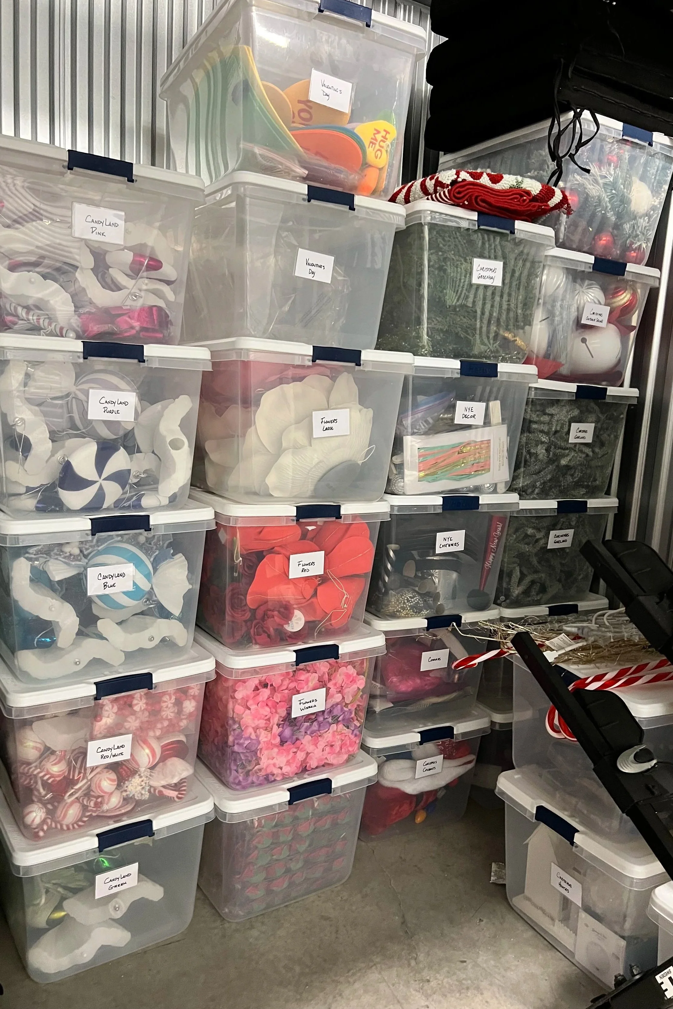 Stacked, labeled clear storage bins of seasonal holiday décor — ornaments, faux flowers, and garland — in an organized storage unit.