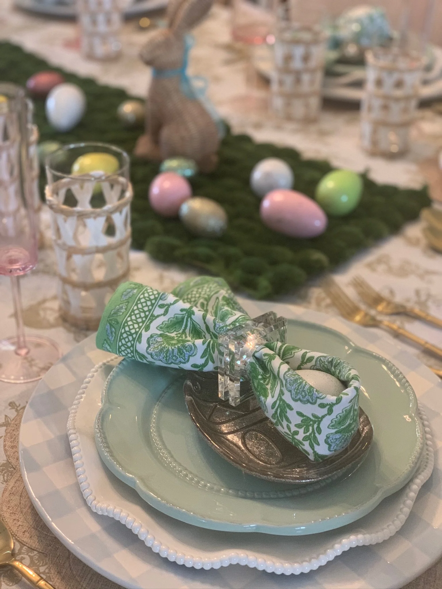 Easter table setting with pastel-colored plates, a green and white patterned napkin, and decorative Easter eggs on a table with a green moss runner and bunny figurine in the background.