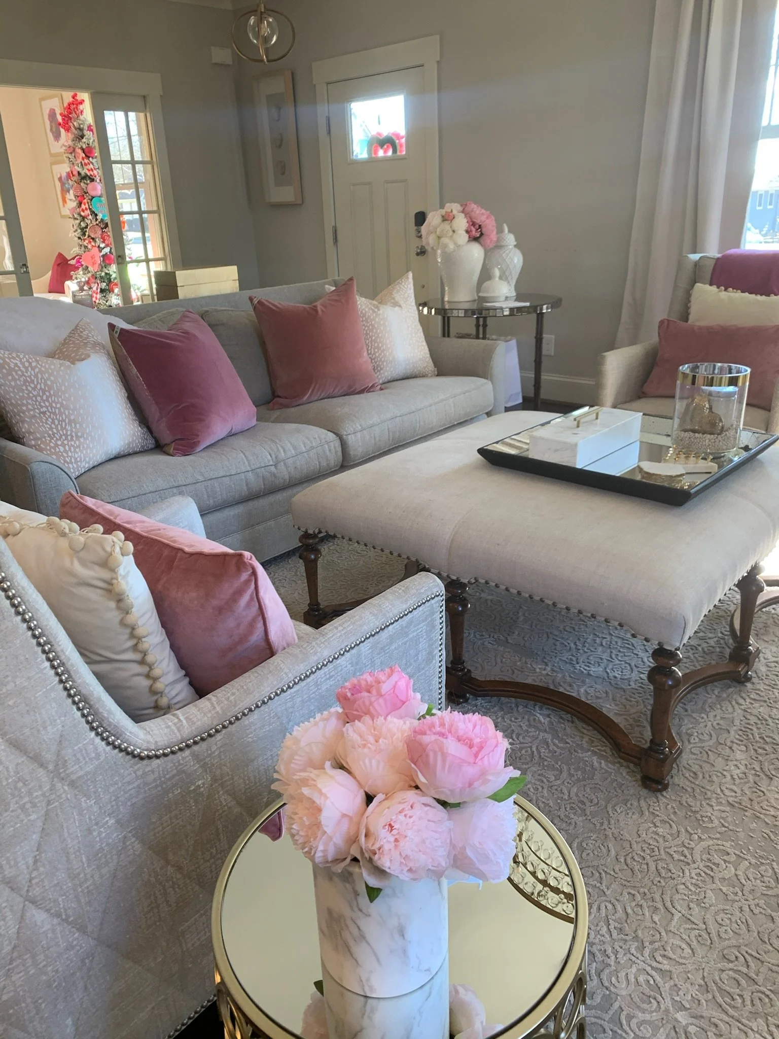 Living room with light-colored sofa and armchair, decorated with pink and cream pillows, a marble vase with pink peonies on a small gold side table, a beige upholstered ottoman in front, a glass tray with decorative items, and a table with white vases and a pink floral arrangement. In the background, a Christmas tree with pink decorations is visible through a window.