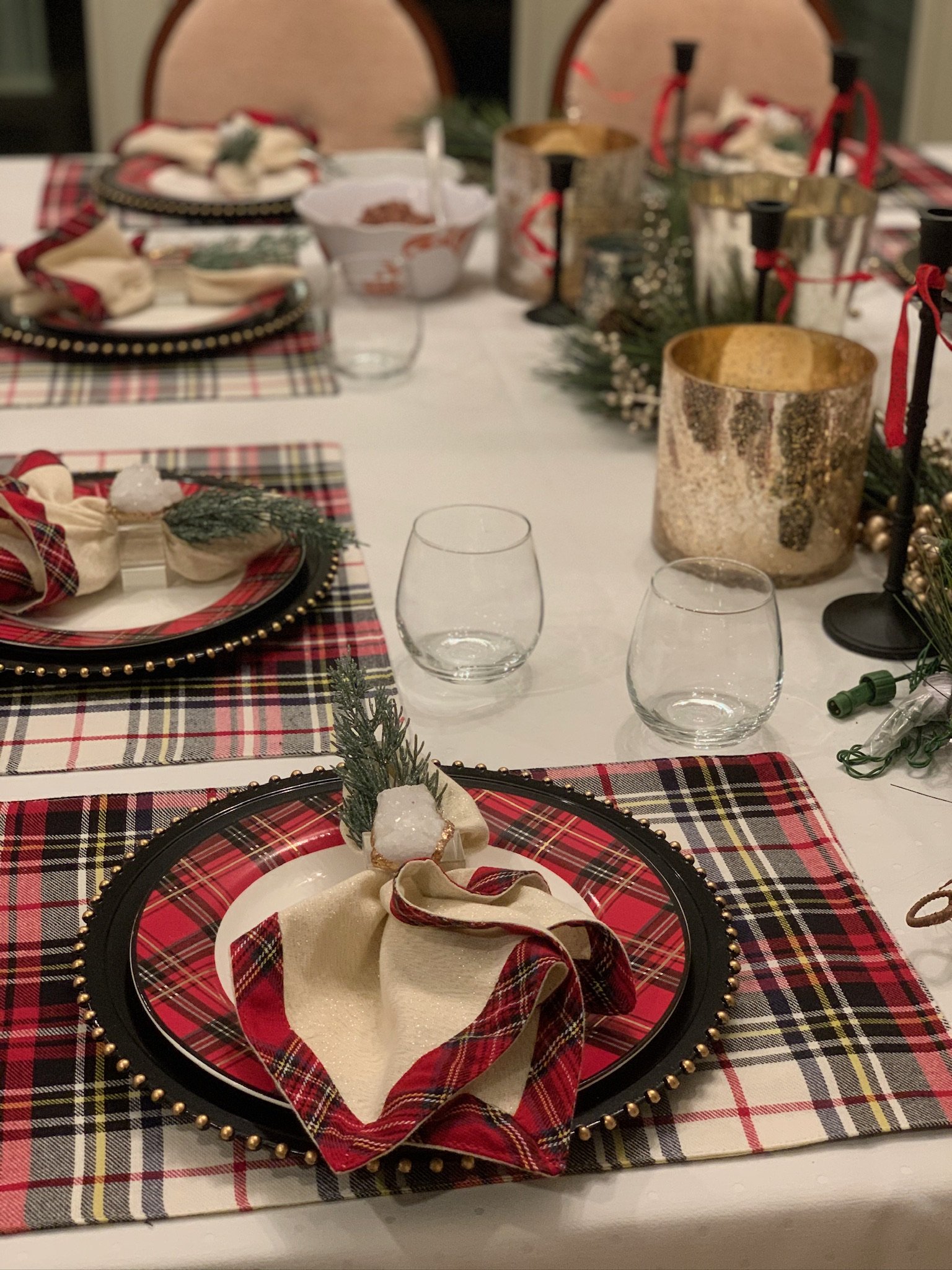 A Christmas table setting with plaid placemats, decorative black plates with gold beads, napkins with checkered patterns, and festive centerpieces including candles, greenery, ornaments, and other decorations.