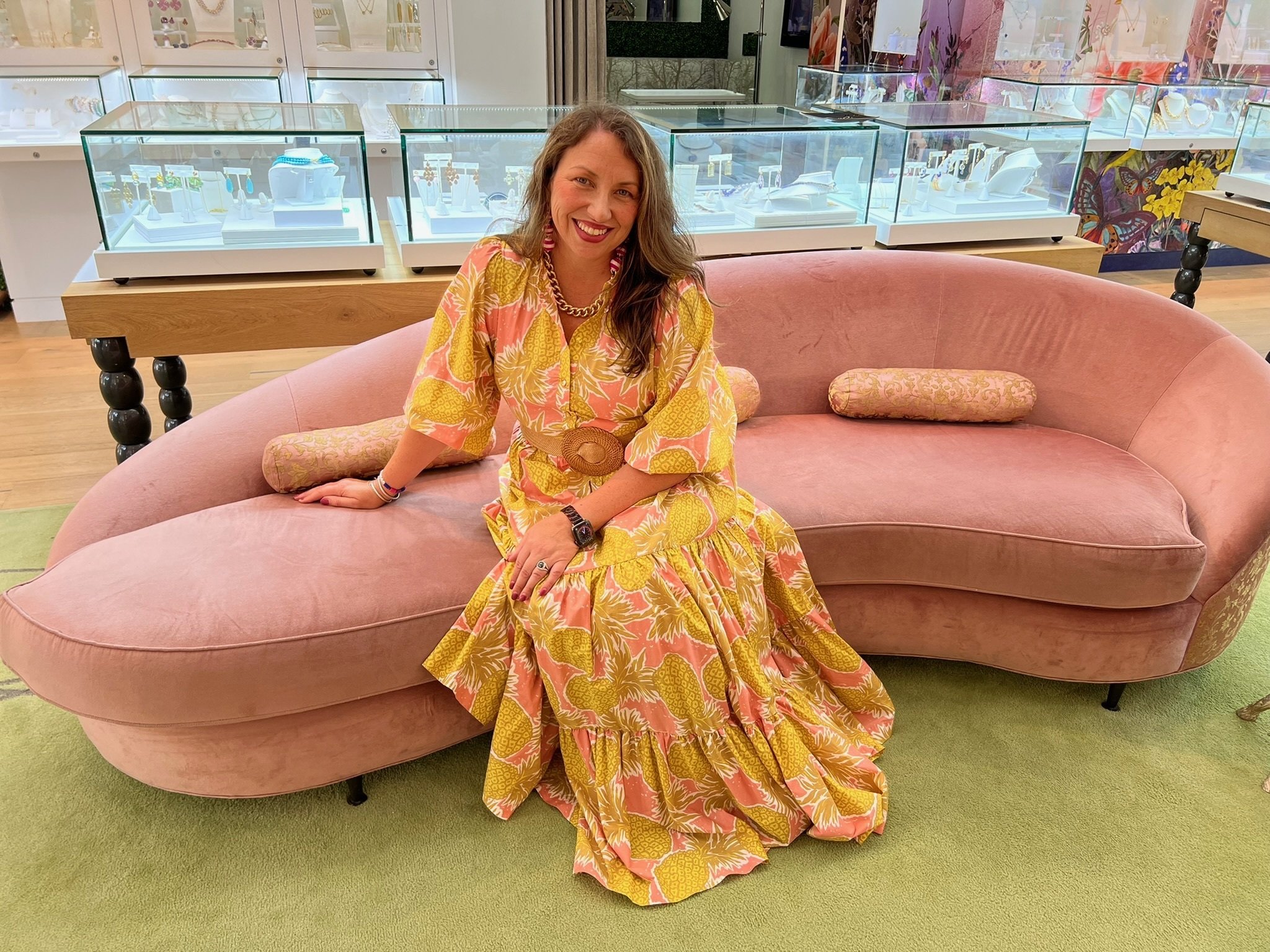 Heather Hagan in a pink-and-yellow printed maxi dress, seated on a curved pink velvet sofa among lit jewelry display cases.