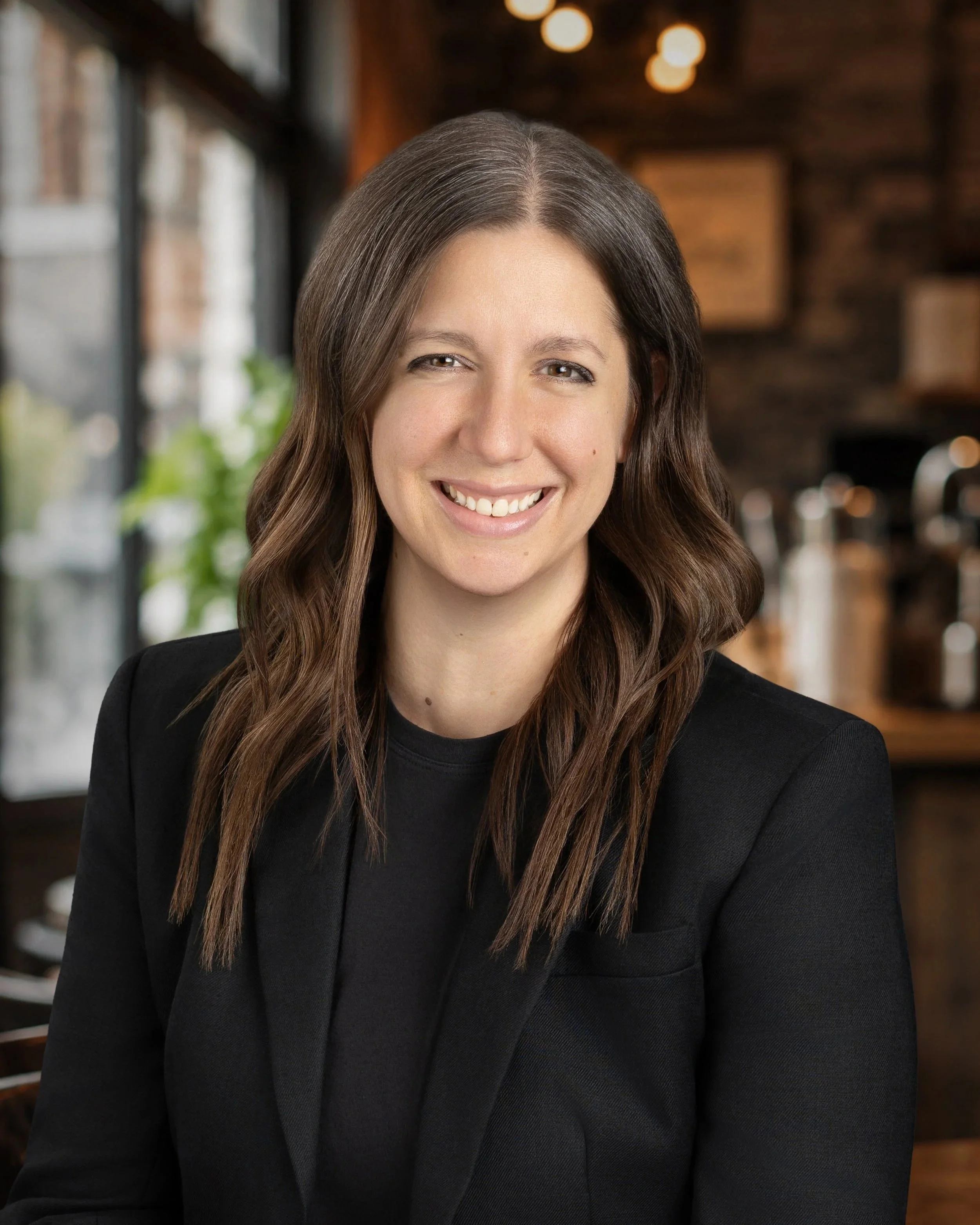 Close-up of a smiling woman with brown hair in loose waves, wearing a black blazer, in a warmly lit indoor setting.