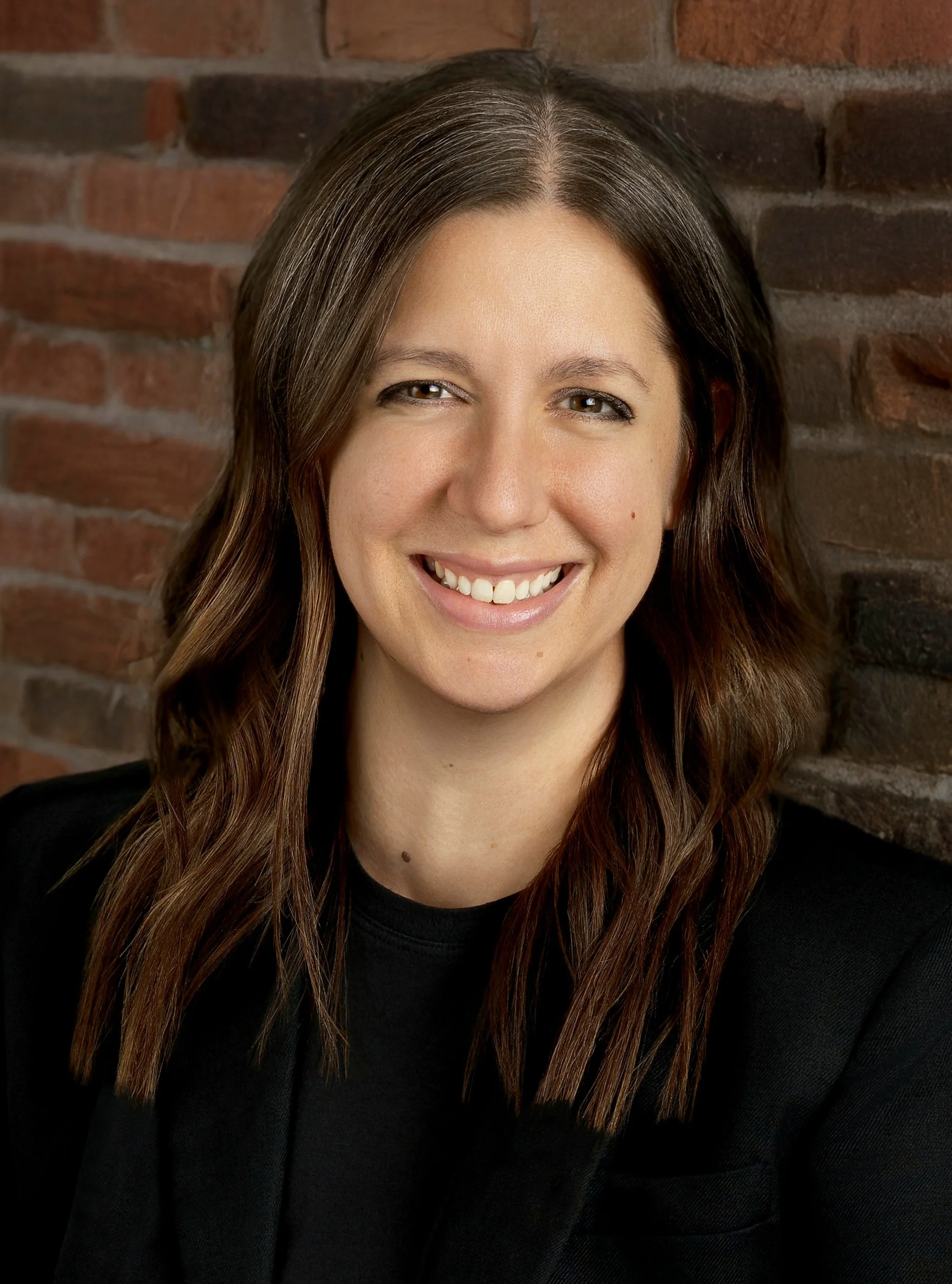 A woman with brown hair, smiling, wearing a black blazer, against a brick wall background.