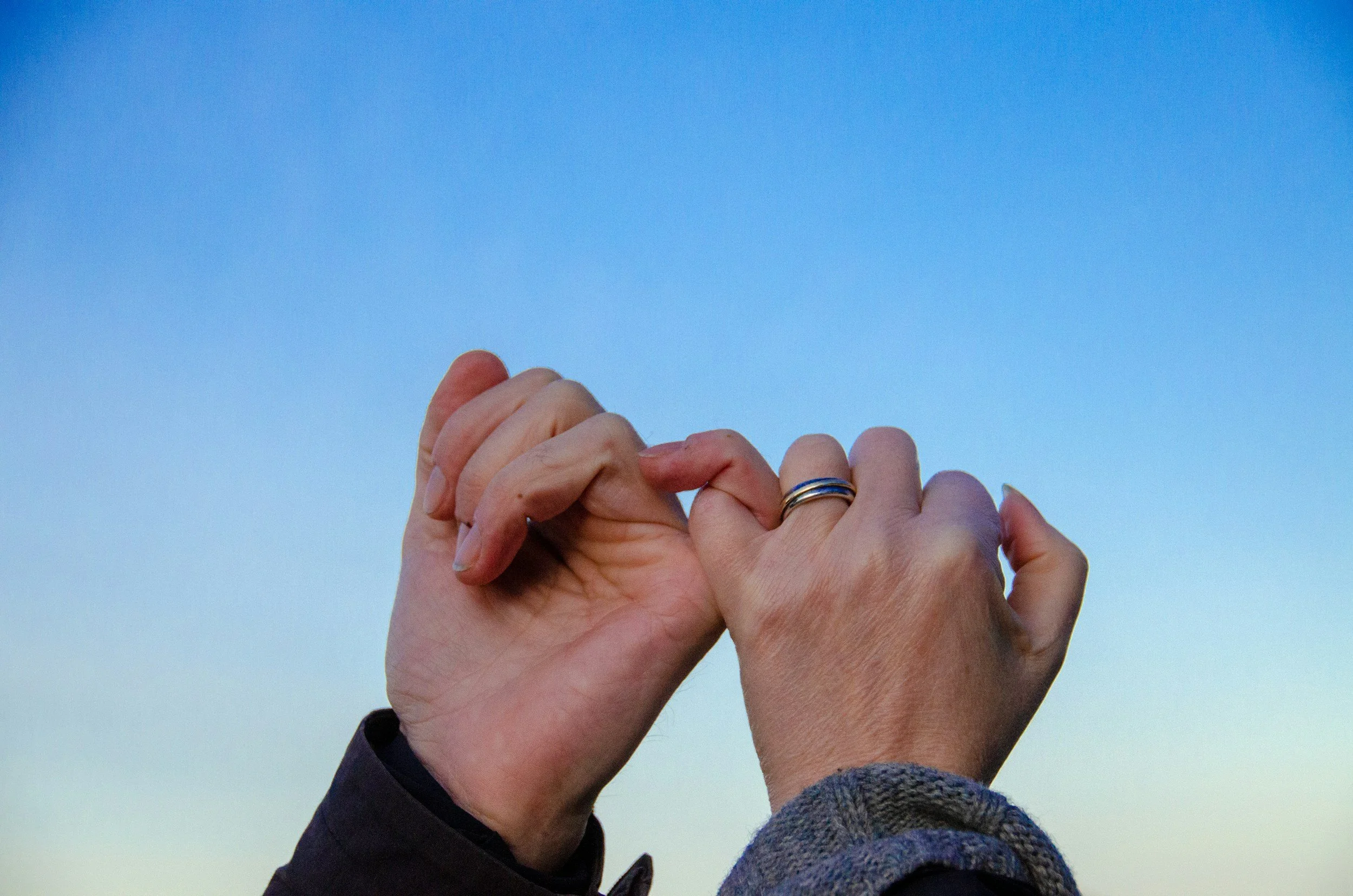 Two hands clasped together against a blue sky, one wearing a ring.