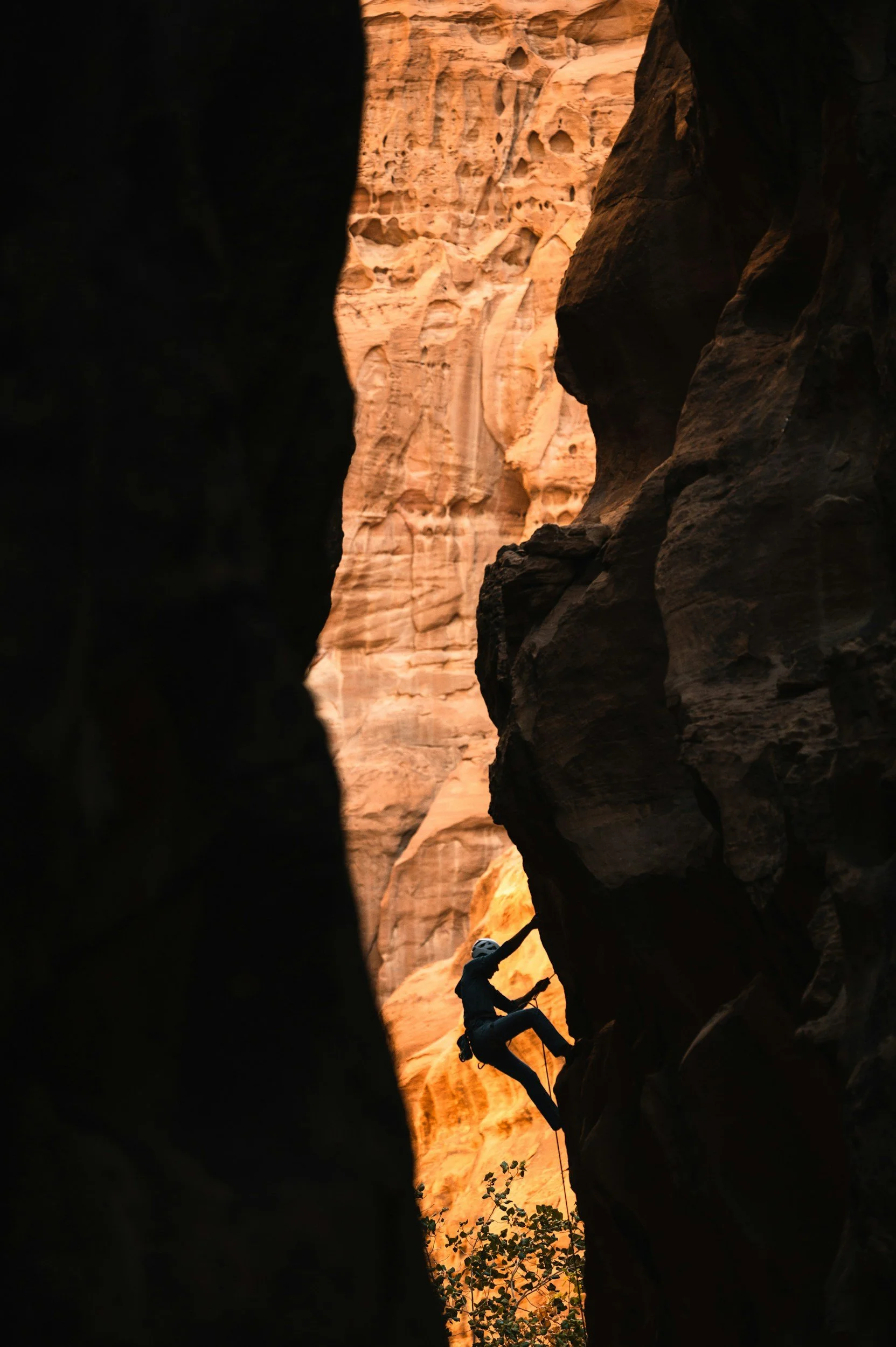 A person wearing a helmet and harness is climbing a rock face inside a narrow canyon with orange rock walls.