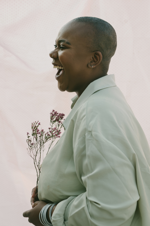 Femme souriante à la tête rasée, portant une veste blanche et tenant un petit bouquet de fleurs sauvages roses et violettes, debout sur un fond clair.