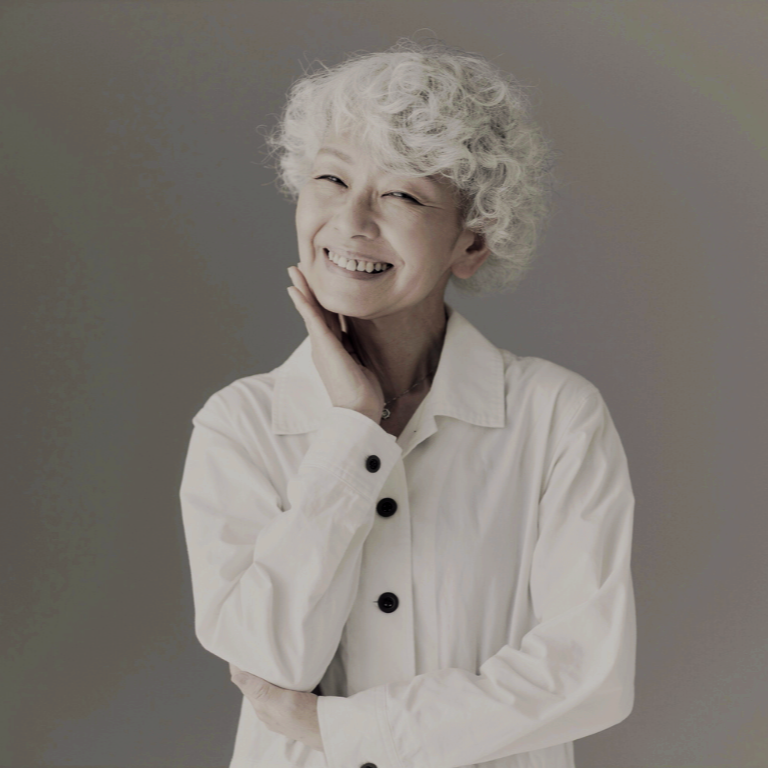 Smiling elderly woman with white curly hair wearing a white button-up shirt, touching her face with one hand.