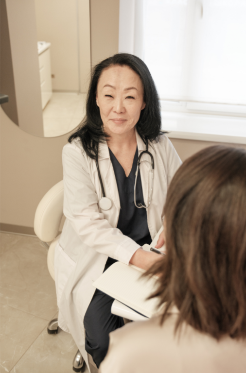 Femme médecin en blouse blanche avec un stéthoscope, assise dans un cabinet médical, parlant avec un patient dont le dos est tourné vers la caméra.