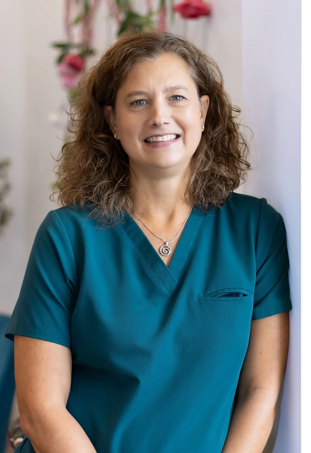 A smiling woman with curly brown hair wearing teal medical scrubs and a silver spiral necklace, leaning against a wall with a blurred background of flowers.