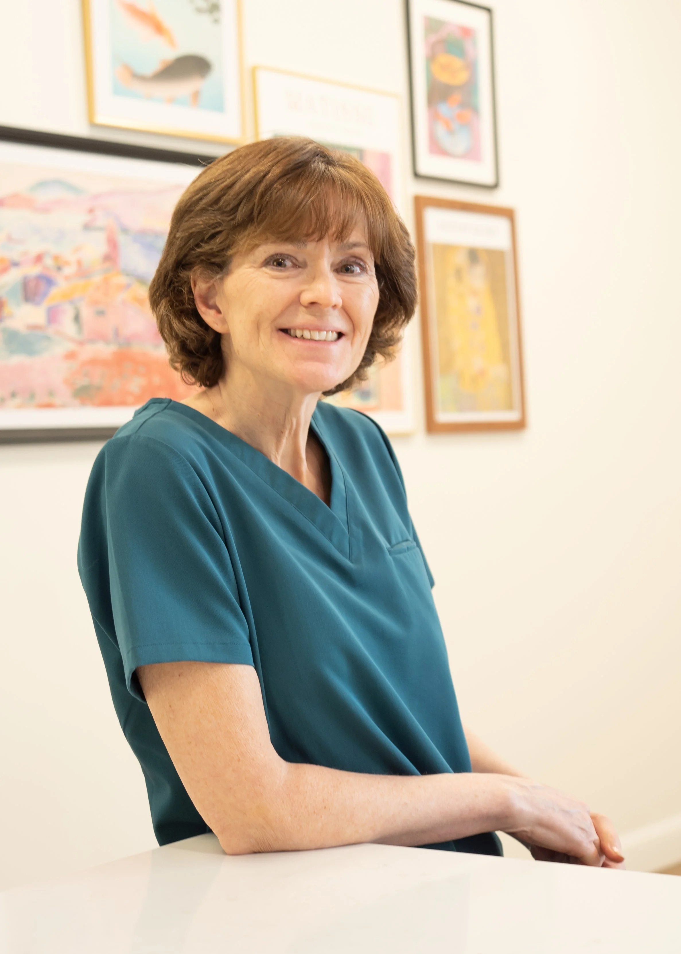 A smiling middle-aged woman with short brown hair wearing a teal medical scrub top, sitting at a white counter with framed colorful artwork on the light-colored wall behind her.