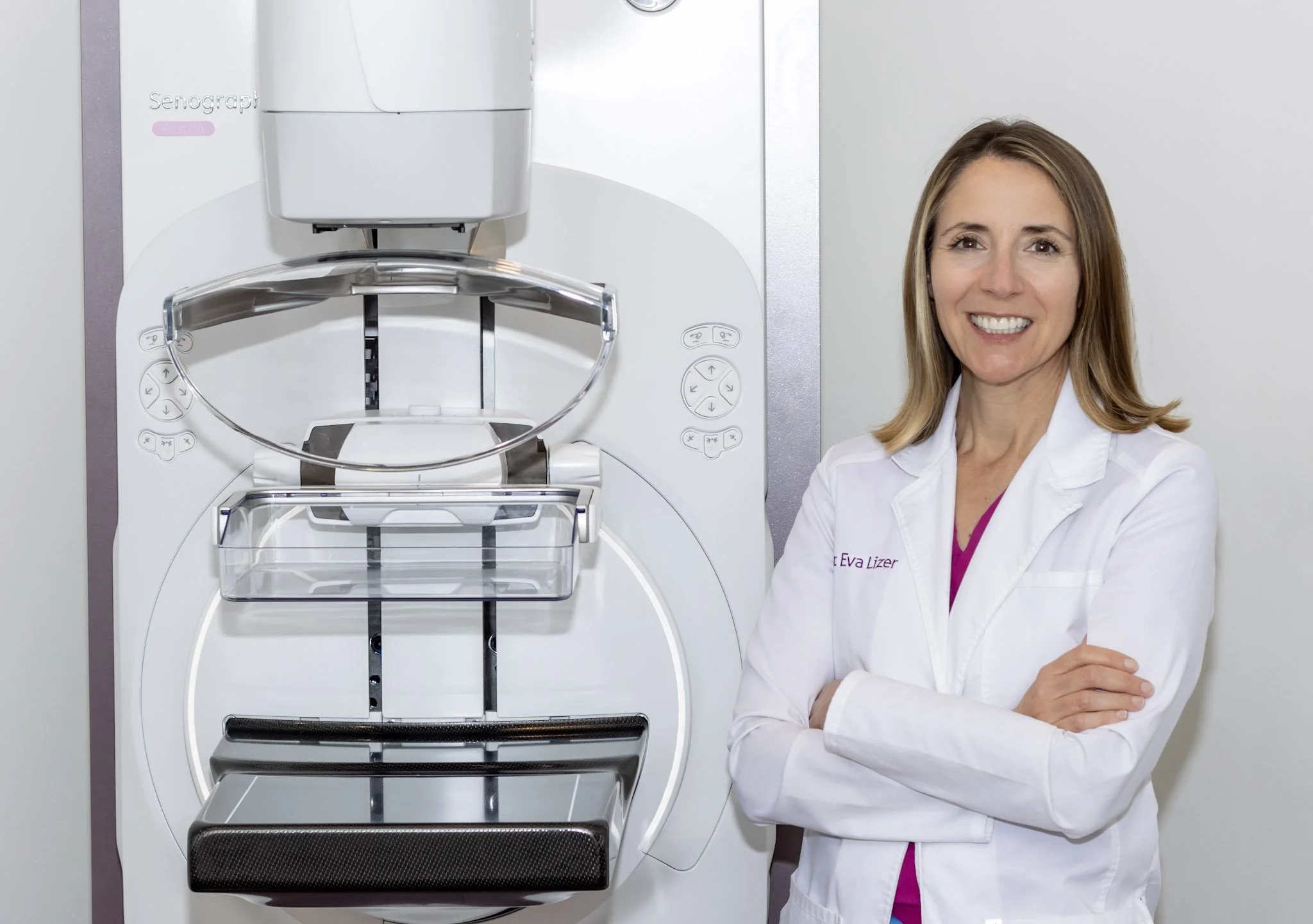 A female medical professional standing next to a mammogram machine, smiling and crossing her arms in a medical setting.