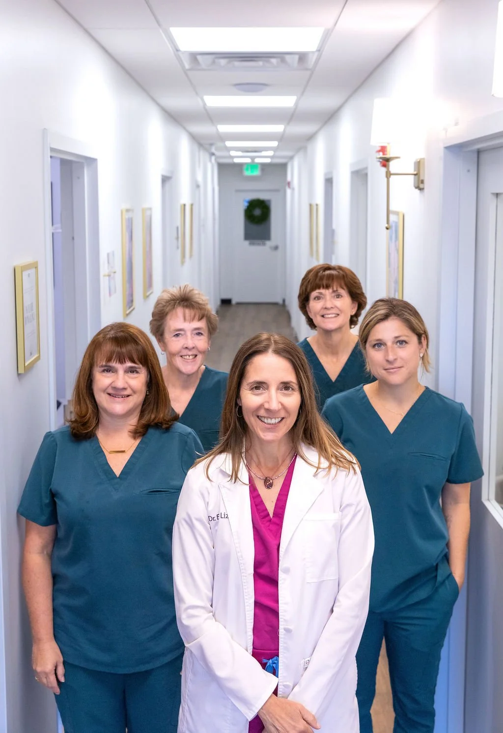 A group of five female healthcare professionals, including a doctor in a white coat, standing in a hospital hallway with white walls, framed pictures, and ceiling lights.