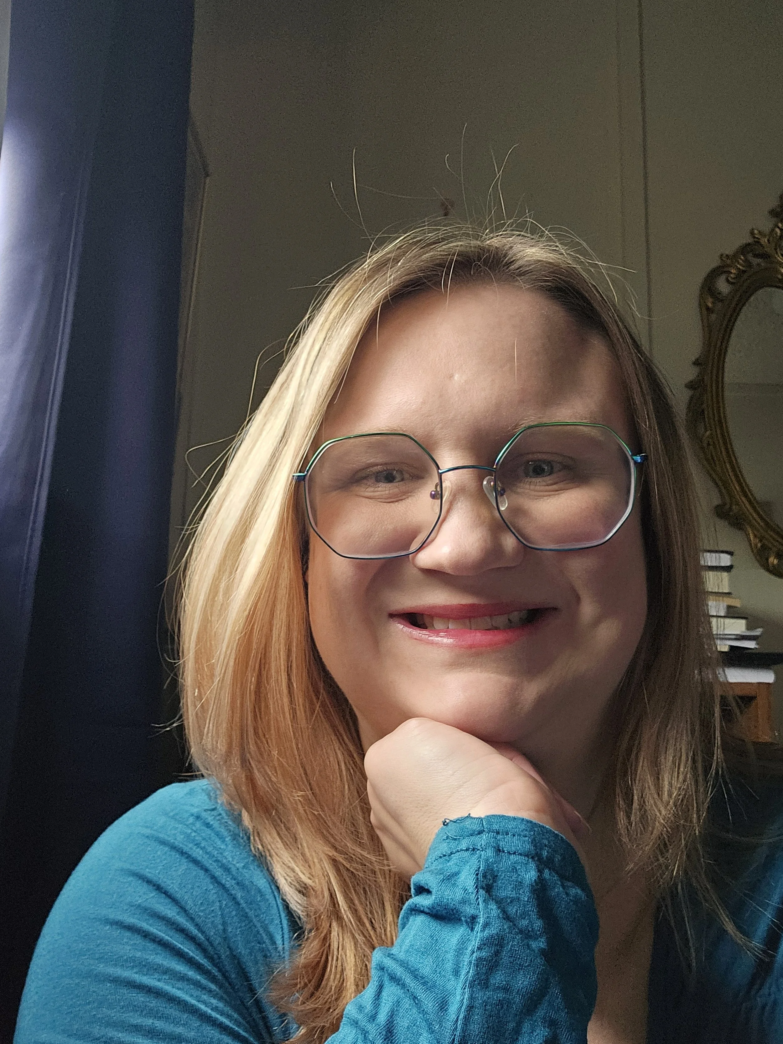 A smiling woman with blonde hair, wearing glasses and a blue top, resting her chin on her hand in a room with books and a decorative mirror in the background.