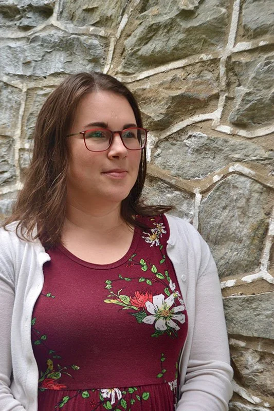 A young woman with shoulder-length brown hair and red glasses standing in front of a stone wall.