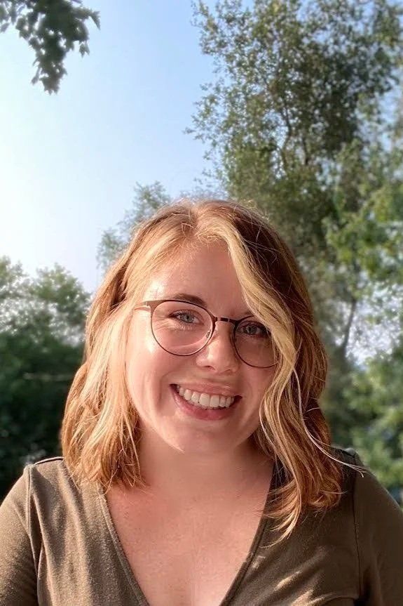 A young woman with shoulder-length black hair and a big smile, wearing a blue blouse, standing in front of green plants indoors.