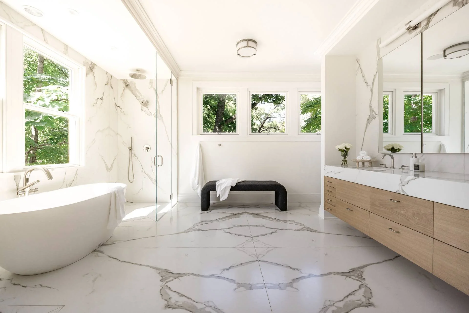 Modern bathroom with white marble floors and walls, a freestanding bathtub, a glass-enclosed shower, a wooden vanity with a large mirror, a black bench, and windows showing green trees outside.