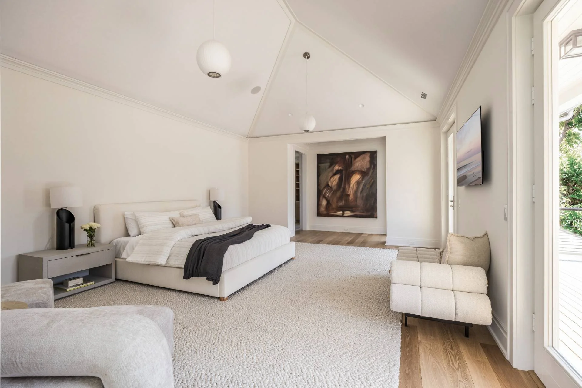 Bright, modern bedroom with white walls, a bed with white bedding and a dark blanket, a nightstand with a small flower, a large wall-mounted TV, a cream-colored bench, and a view of greenery outside through open doors.