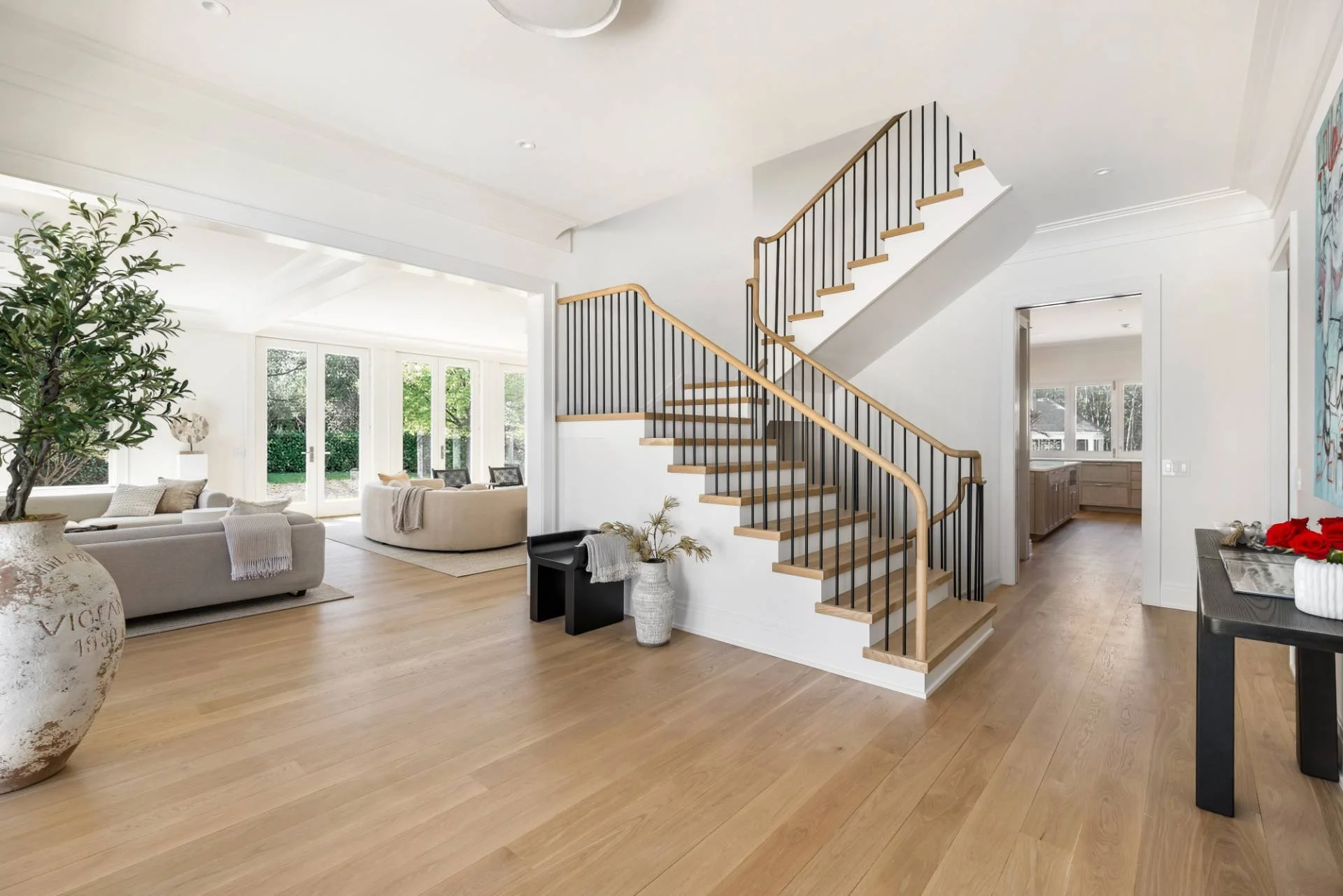 Bright living room with wooden floors, a staircase with black iron railing and wooden handrail, large glass doors leading outside, white walls, and modern furniture.